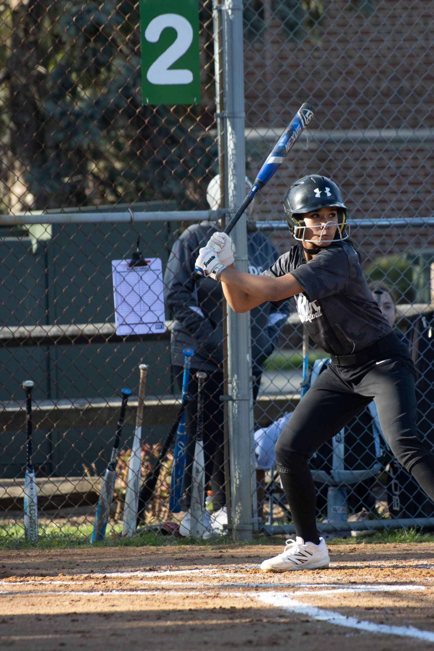 A softball player is standing at the home plate, ready to hit the ball.