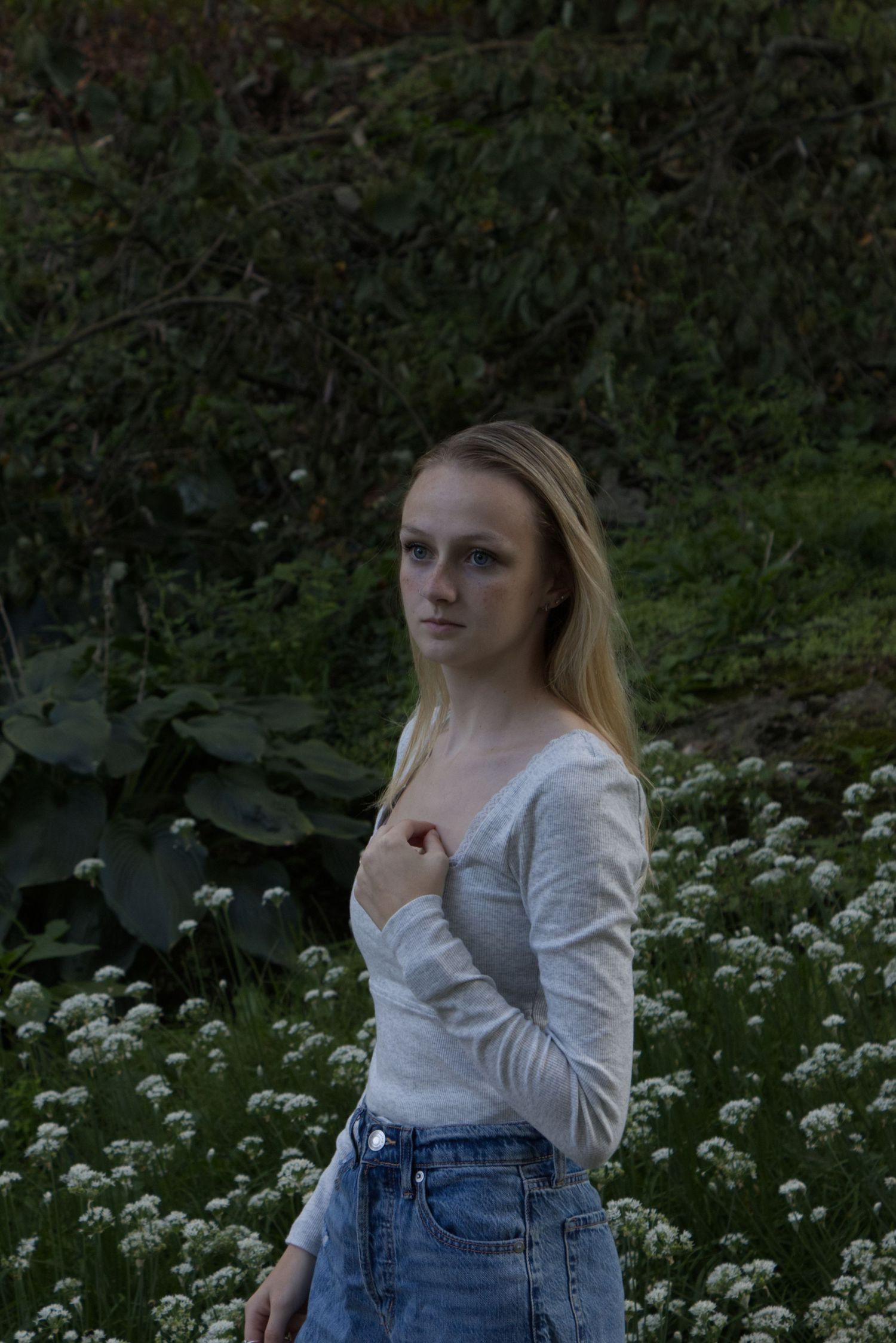 A young woman is at the center of the frame, wearing a plain white shirt, and plants are visible in the background.