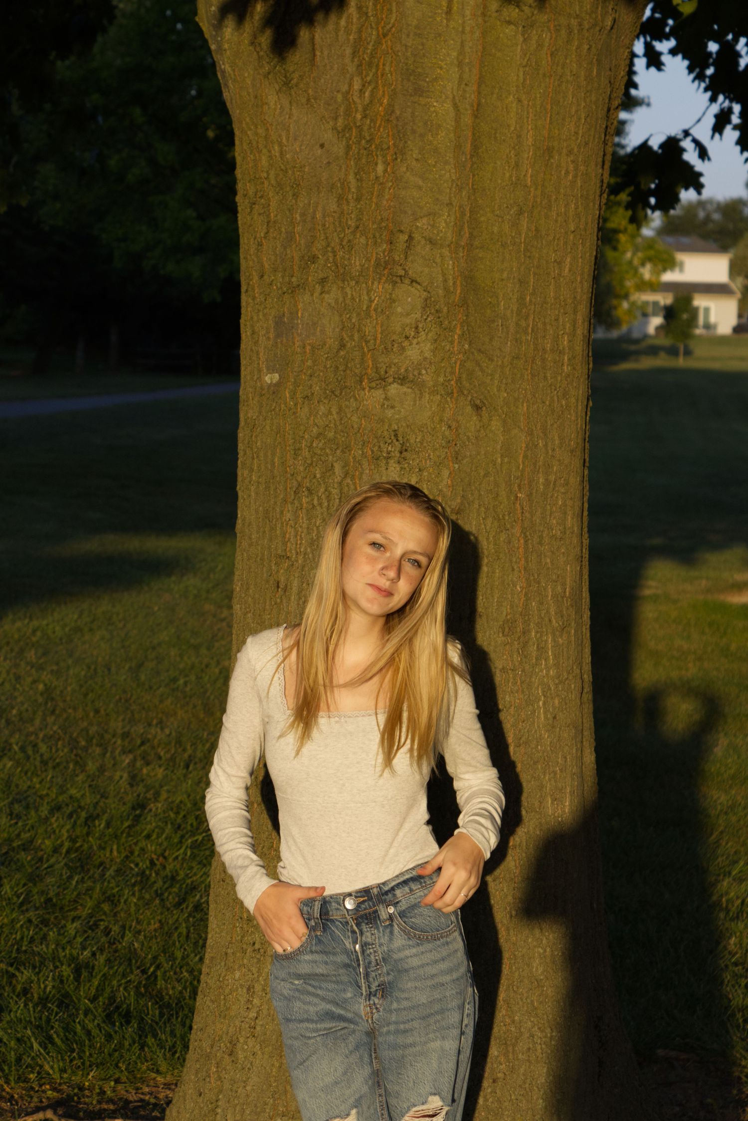 A young woman in a plain white shirt stands against a tree, bathed in the golden hour's lighting.