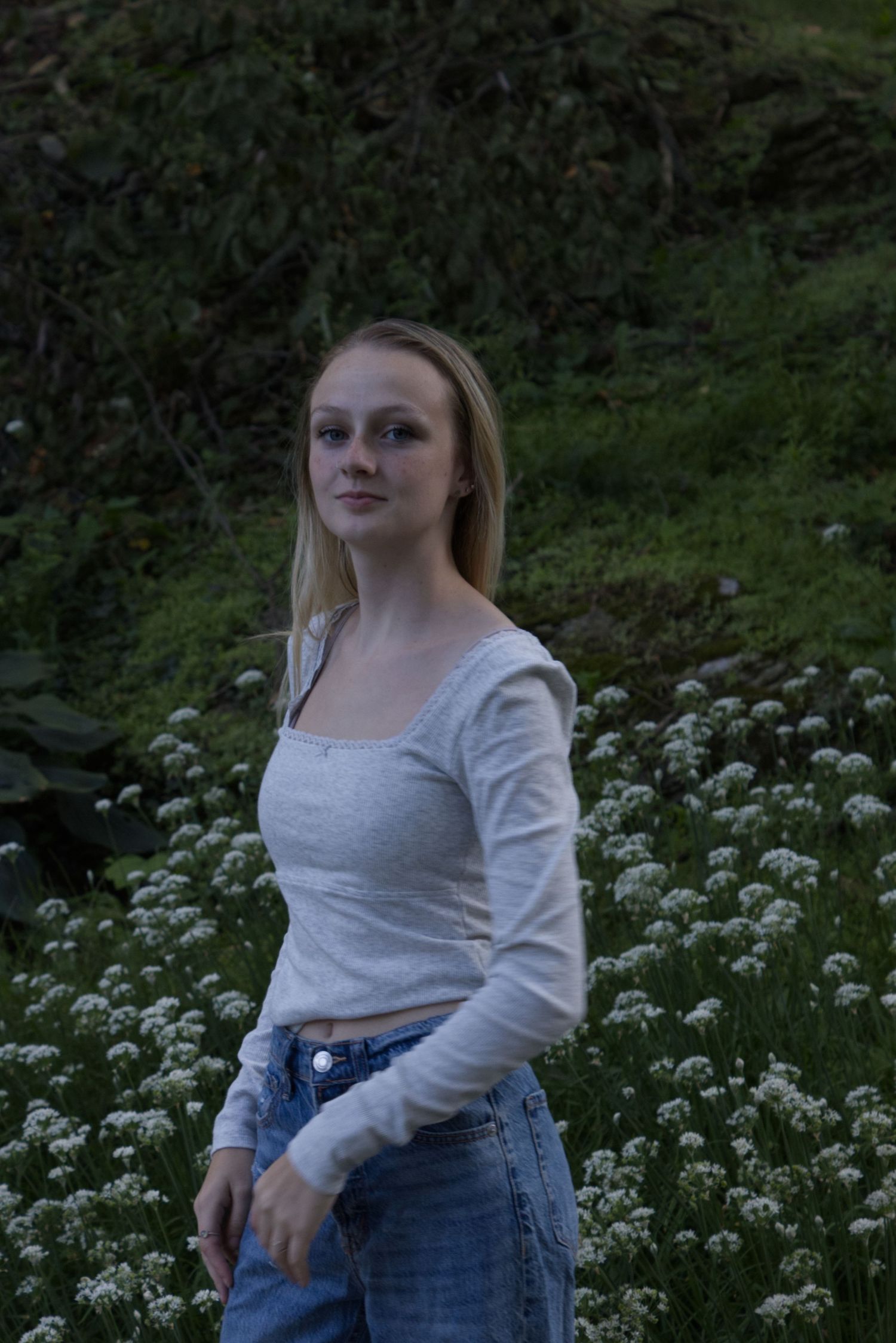 A young woman is at the center of the frame, wearing a plain white shirt, and plants are visible in the background.