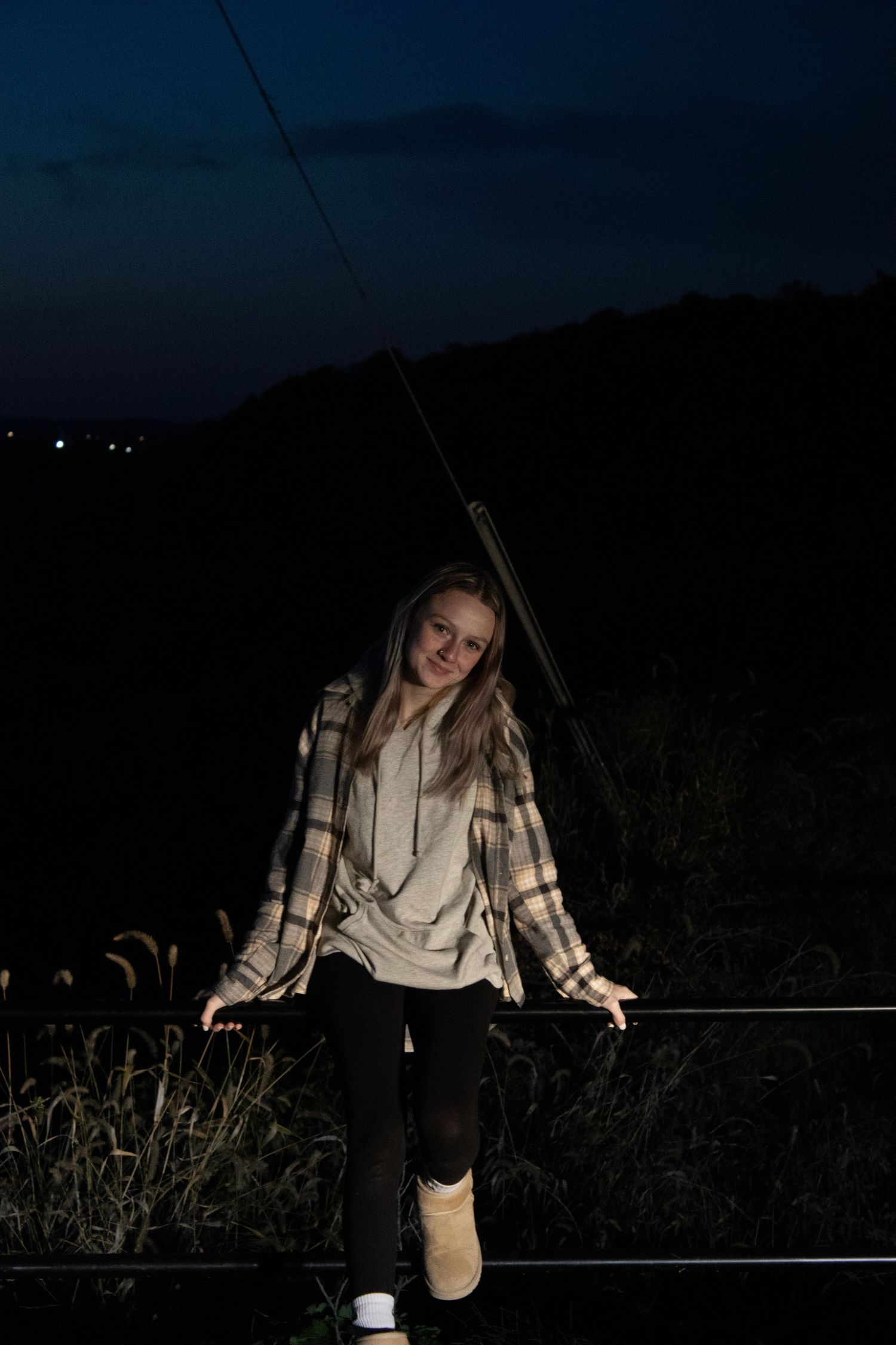 Young woman in a plaid shirt and boots standing on a fence rail at night, softly lit against a dark hillside.