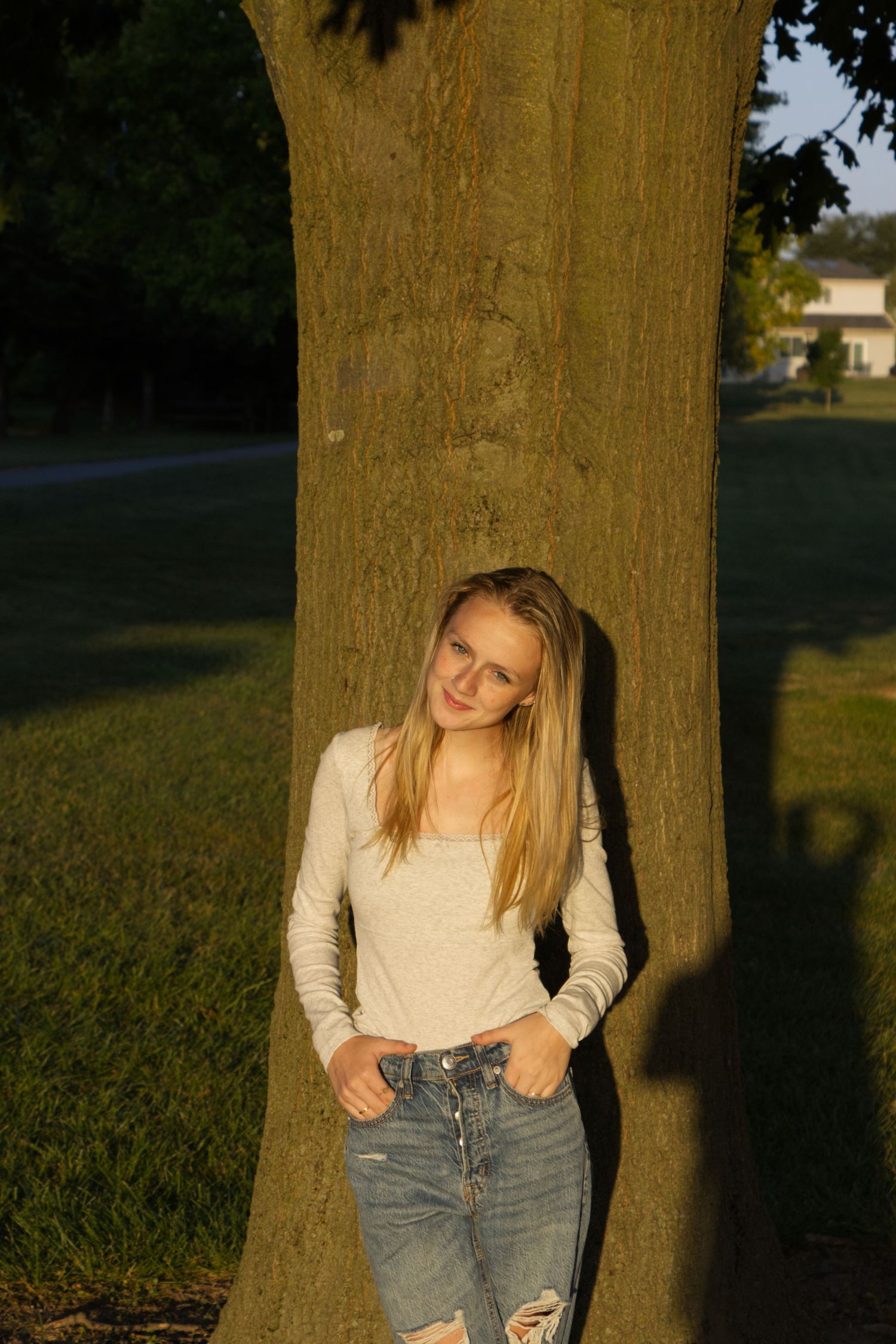 A young woman in a plain white shirt stands against a tree, bathed in the golden hour's lighting.