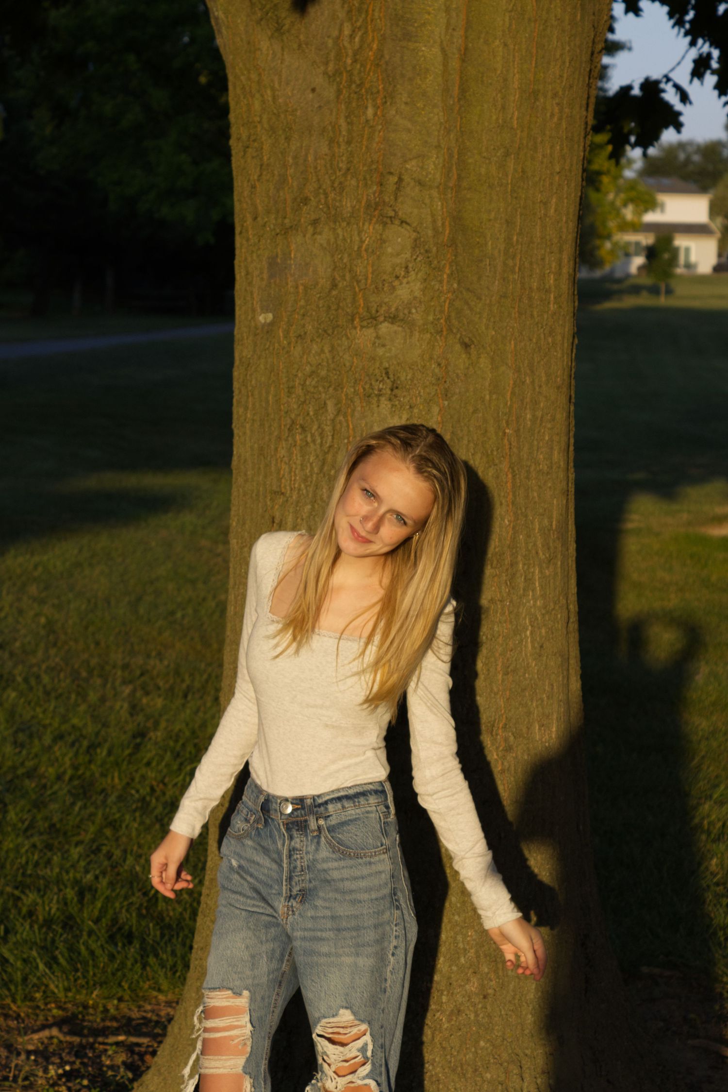 A young woman in a plain white shirt stands against a tree, bathed in the golden hour's light.