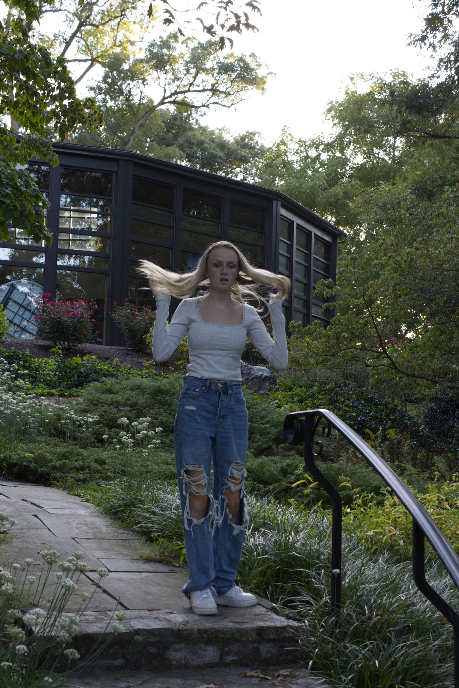 A young woman, standing on a set of stairs, is gazing directly at the camera while also flicking her hair.