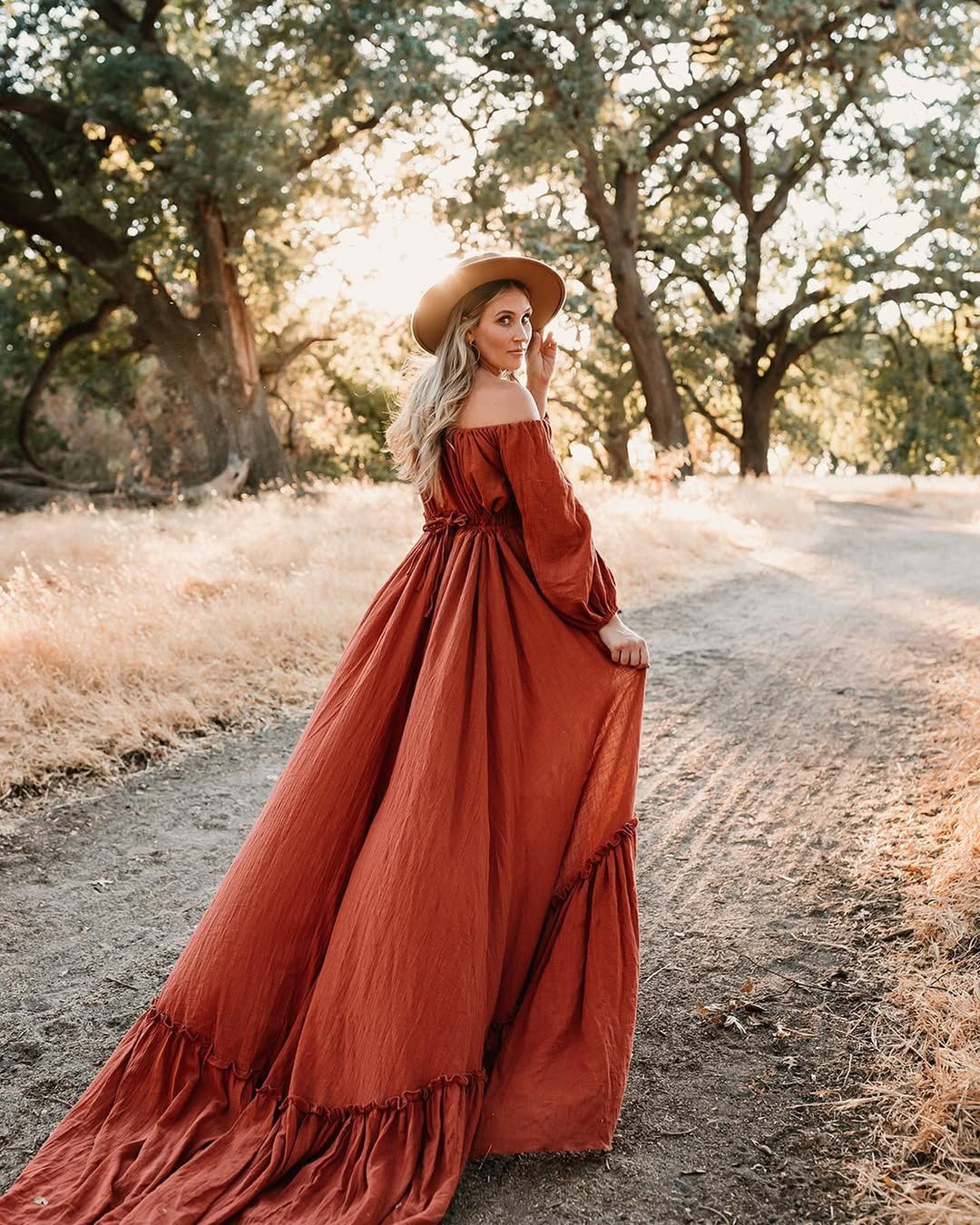 Long flowing rust red dress photographed on dirt path with golden sunset through trees.