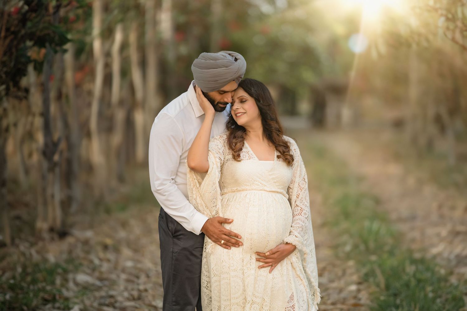 Intimate maternity moment captured in golden sunset light through wooded area.