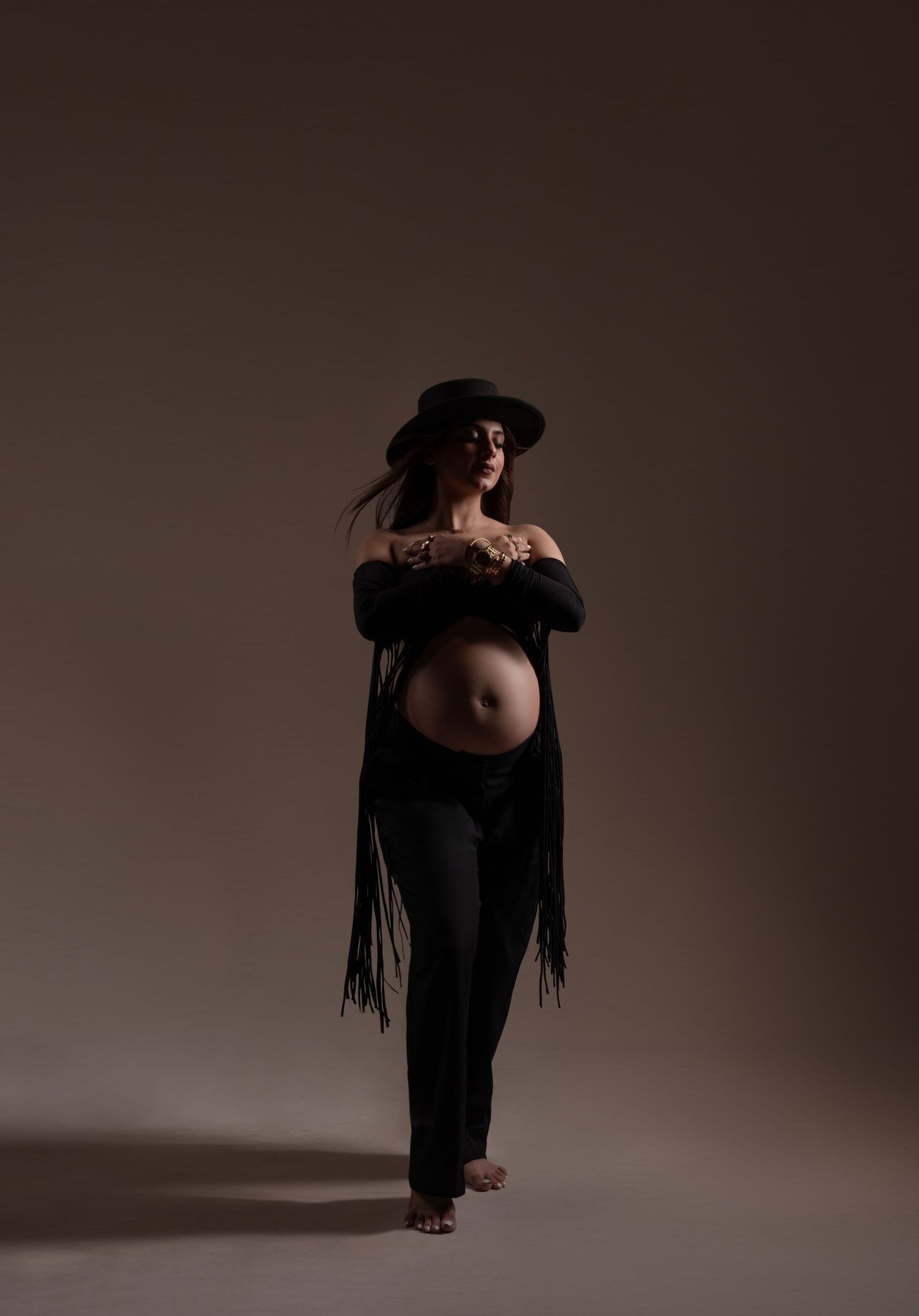 Dramatic maternity portrait in black fringe coat and hat against dark background.