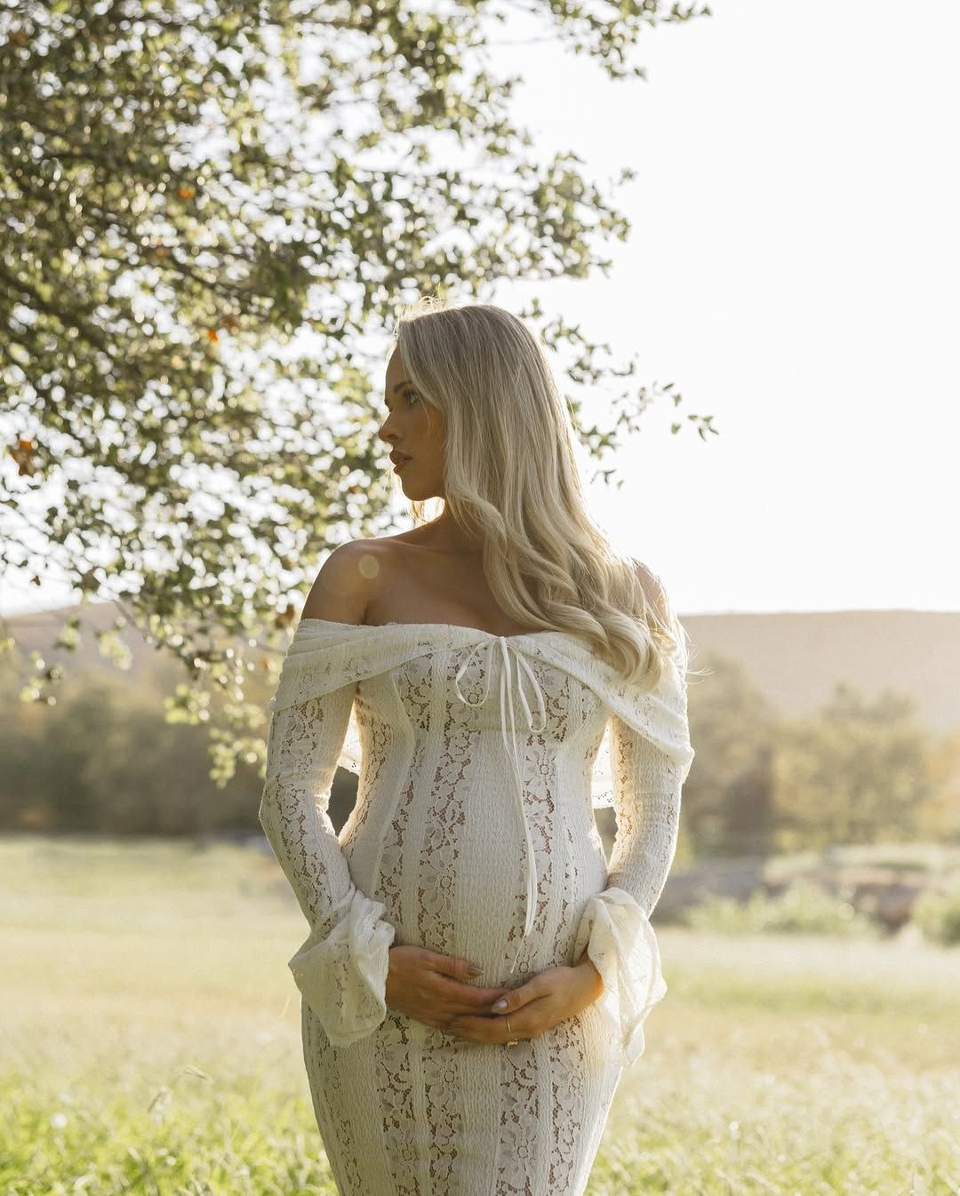 Backlit maternity portrait in lace dress during golden hour in countryside setting.