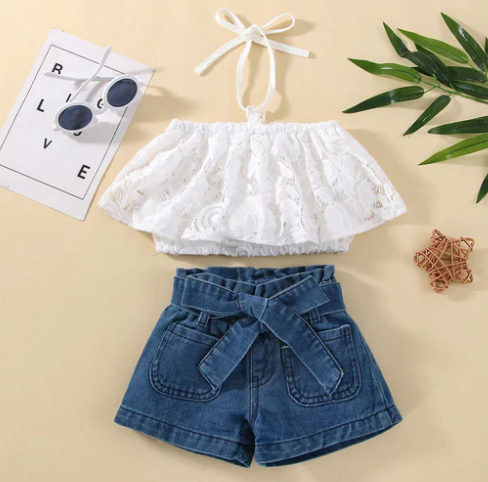 Delicate white lace baby romper with ruffle details and tie waist displayed with floral accents.