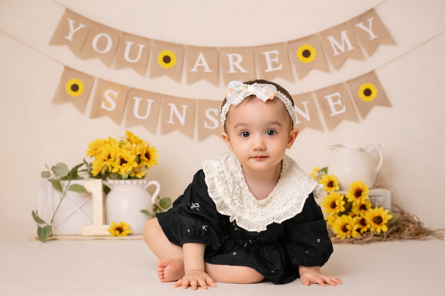 A baby in a black dress and white headband sits in front of a 