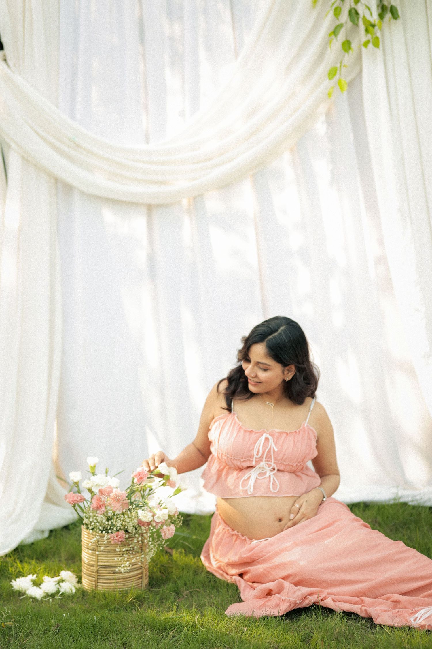 Pregnant woman in pink crop top and skirt sits on grass touching flowers near white draped backdrop.