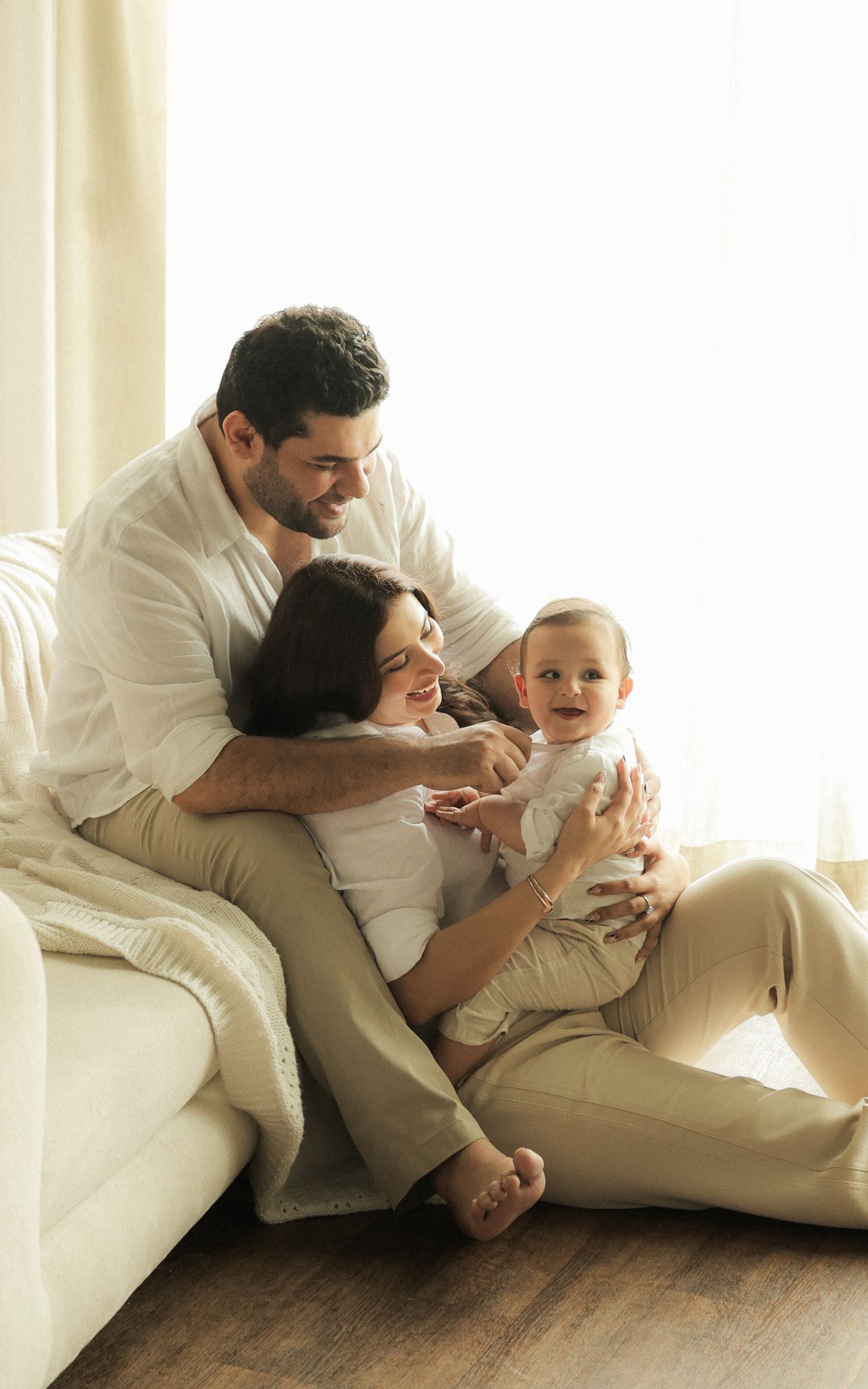 A happy young family with a smiling baby sits together on the floor by a couch in a bright, airy living room.