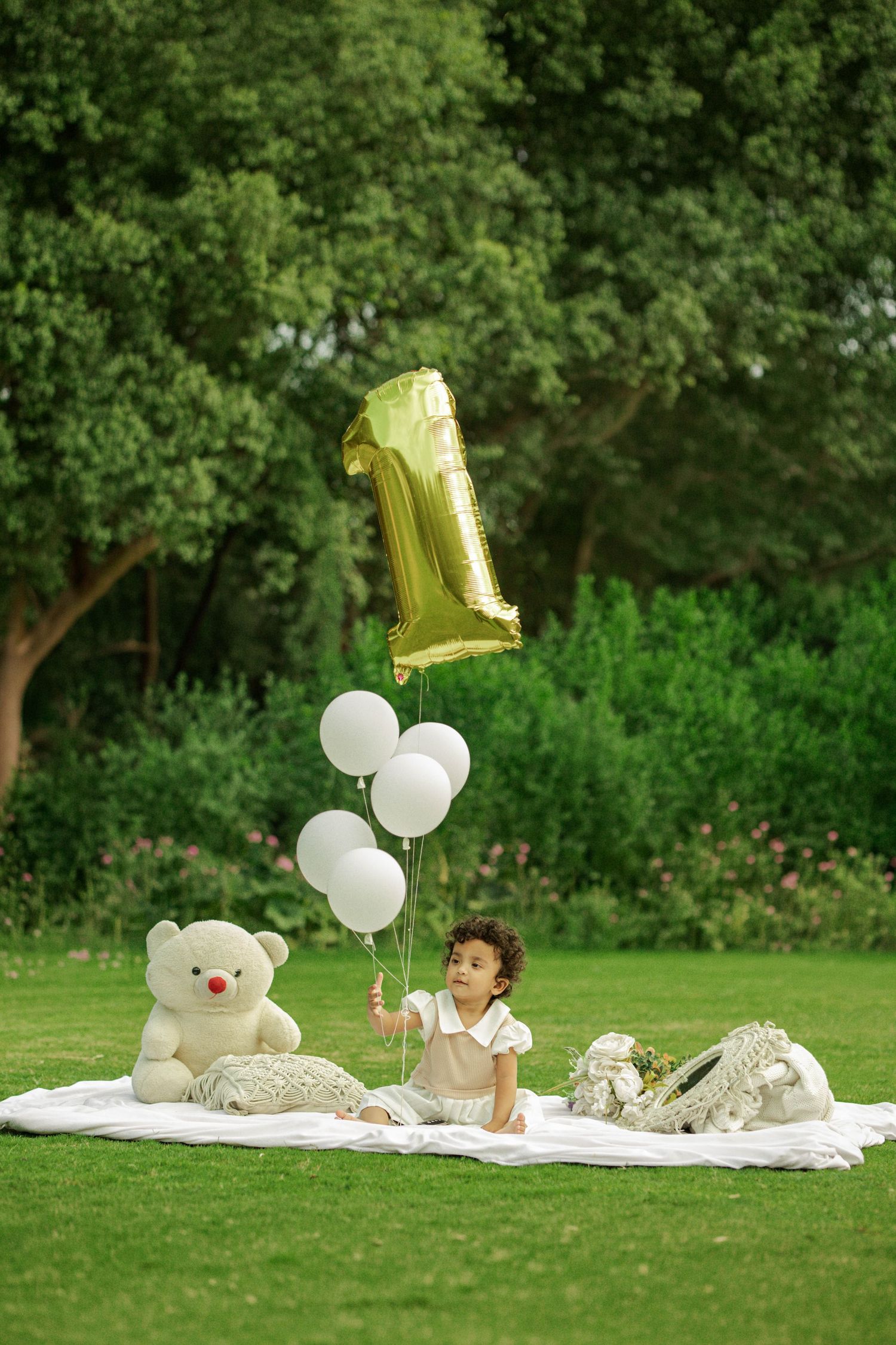 Baby's first birthday photoshoot on grass with gold number 1 balloon, white balloons, and teddy bear on white blanket.