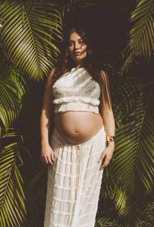 Maternity photo featuring a white lace two-piece outfit against tropical palm fronds in moody lighting.