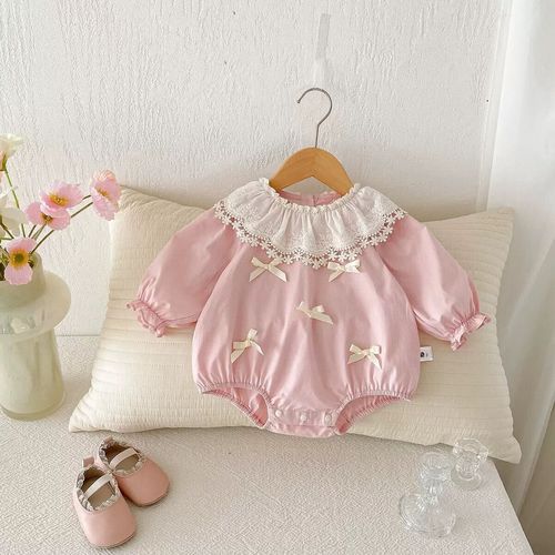 Vintage-style pink baby romper with white lace collar and bows displayed with matching pink shoes on white pillow.