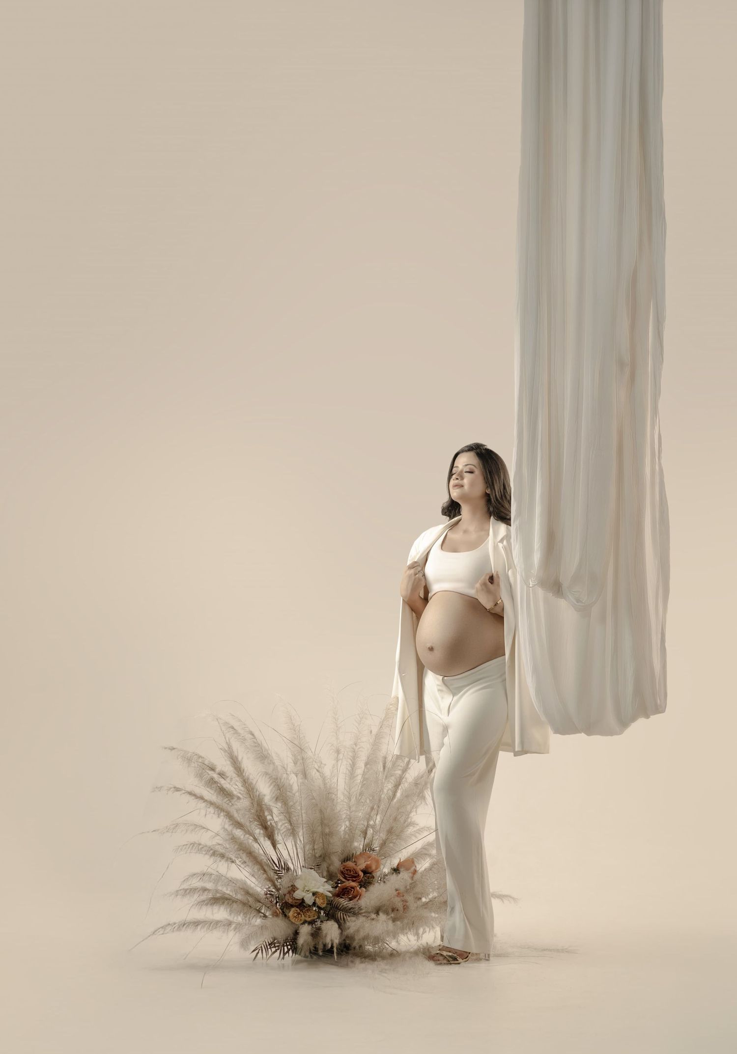 Artistic maternityportraits of a figure in white flowing fabric against a light backdrop with dried pampas grass arrangements.