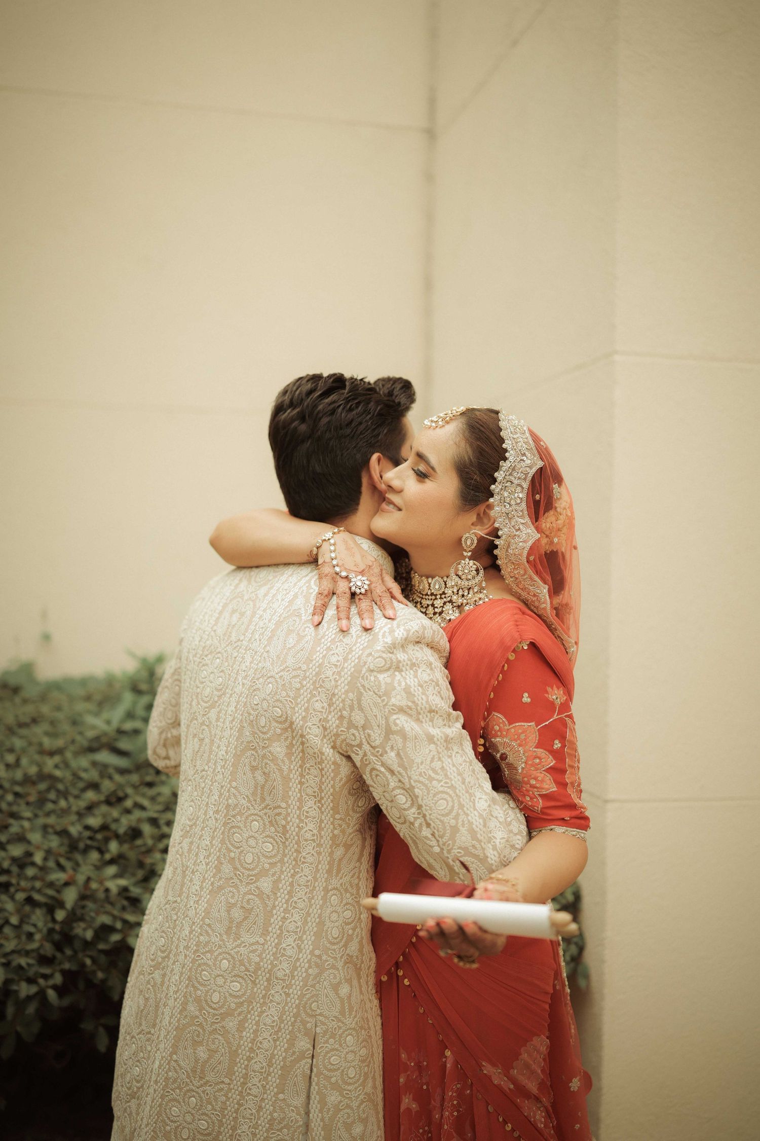 Indian bride in red saree and groom in white sherwani share a romantic embrace during traditional wedding ceremony.