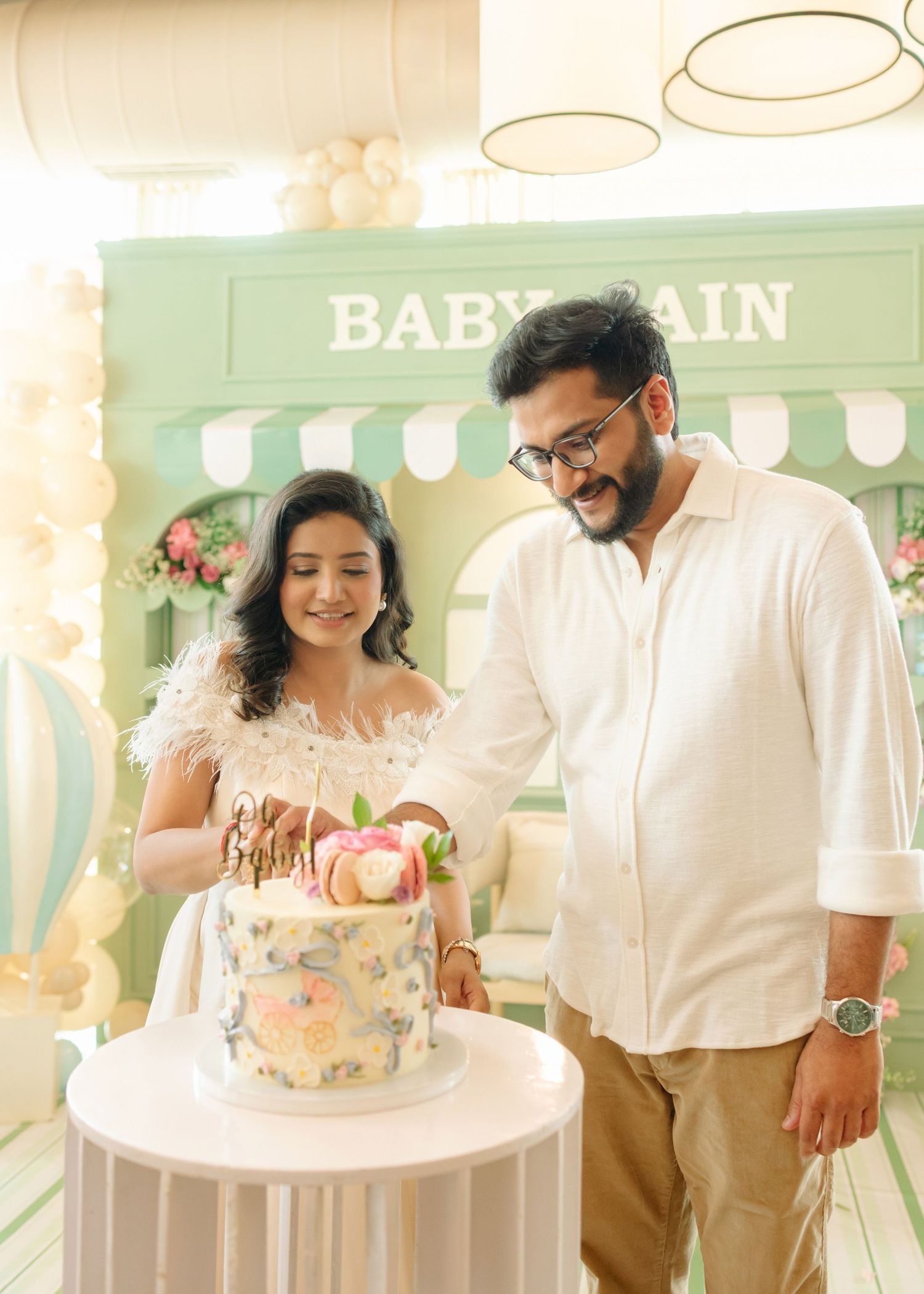 Couple cutting a decorated cake at a baby shower party with a mint green backdrop.
