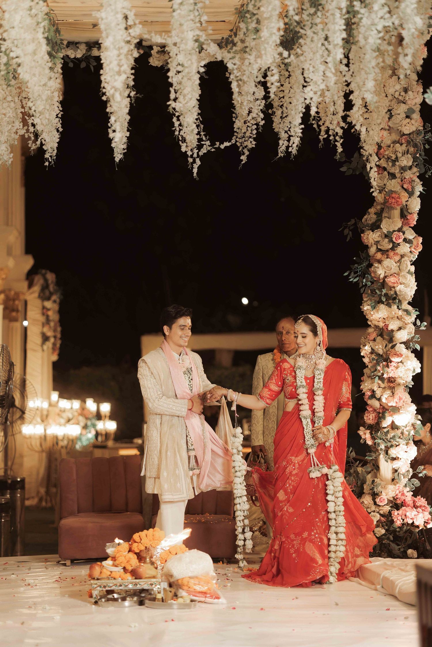 Indian bride in red saree and groom in sherwani exchange garlands at floral-decorated wedding ceremony.