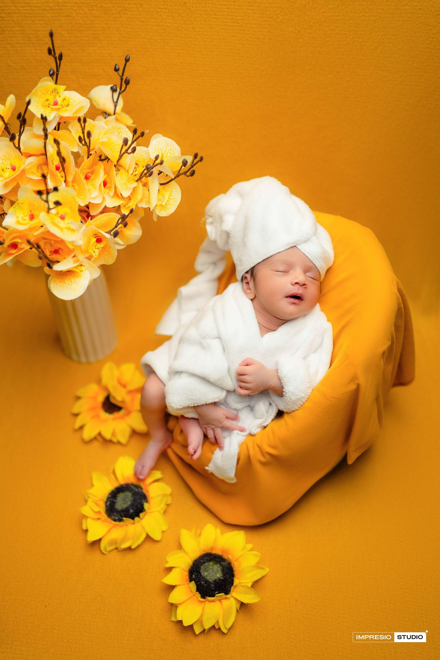 Newborn baby sleeping in white robe and towel hat on yellow mini sofa surrounded by sunflowers.