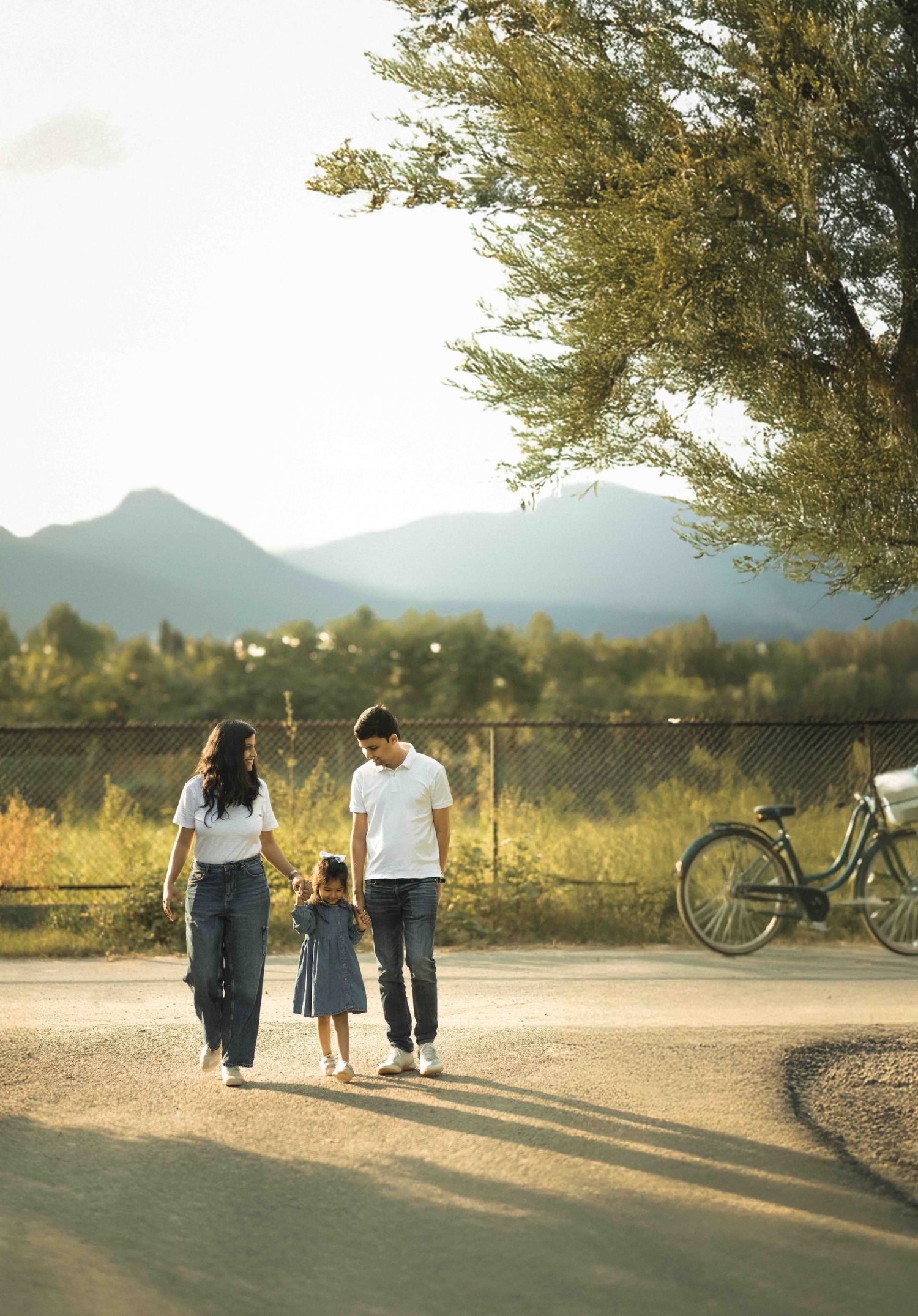 Family of three walking hand-in-hand outdoors near mountains, with a bicycle and trees in warm golden sunlight.