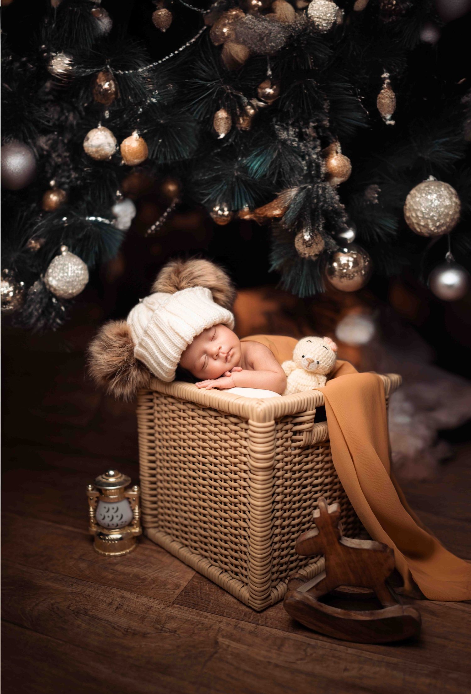 Newborn baby sleeping in wicker basket wearing white fur hat under Christmas tree with gold ornaments.