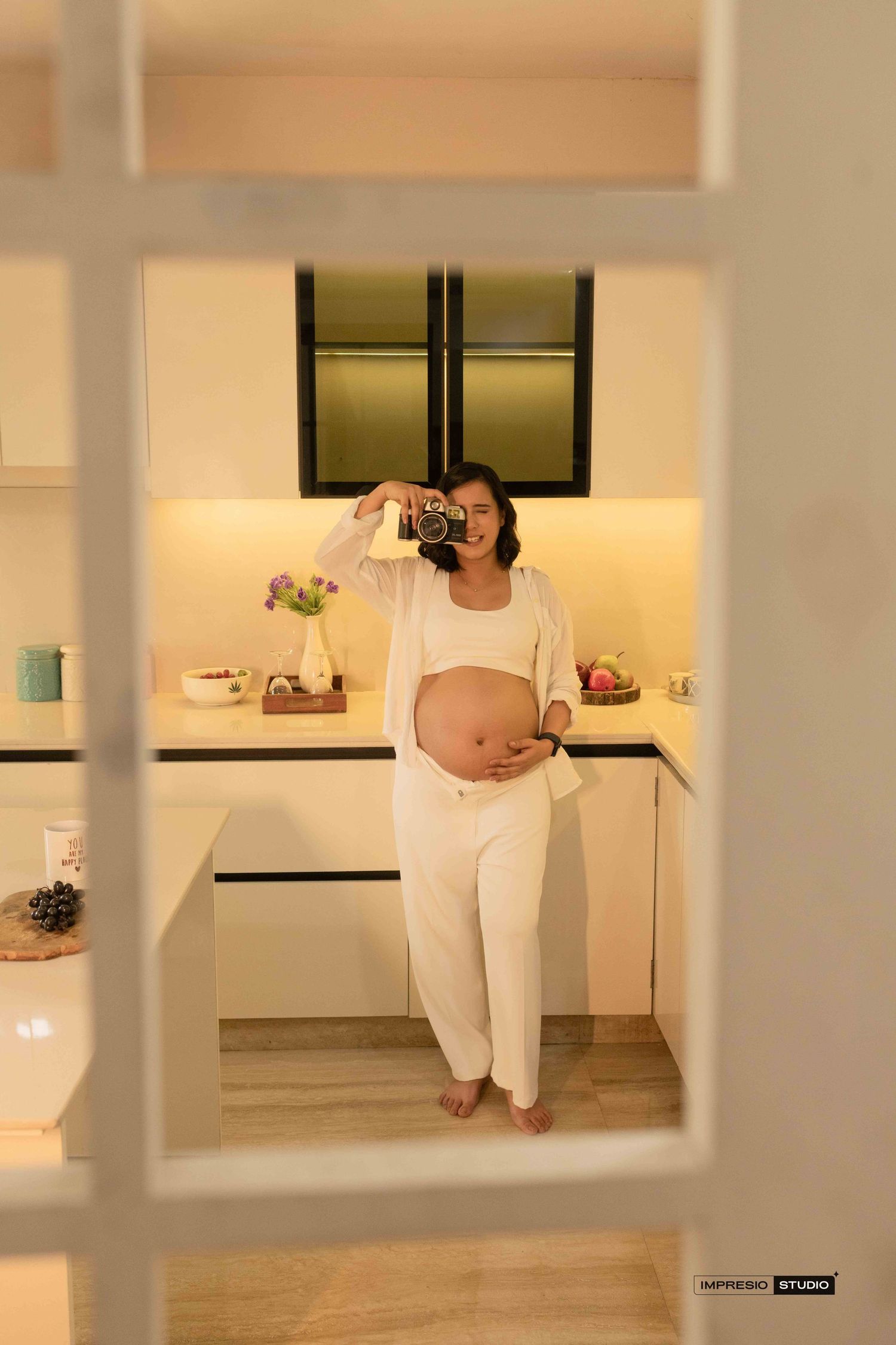Pregnant woman in white outfit holding camera in modern kitchen, maternity photography session.