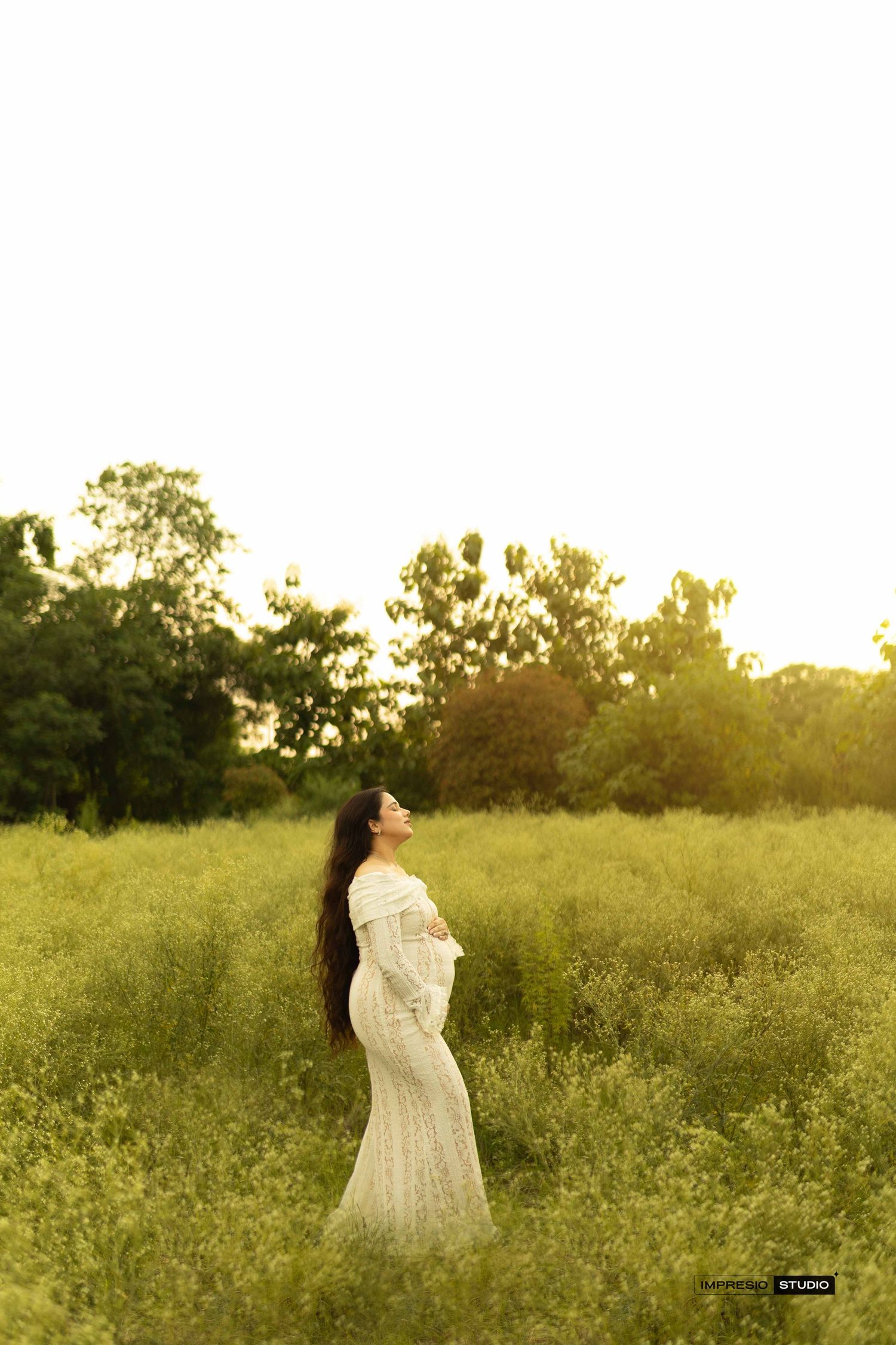 Pregnant woman in white lace dress standing in green field at golden hour, cradling baby bump.