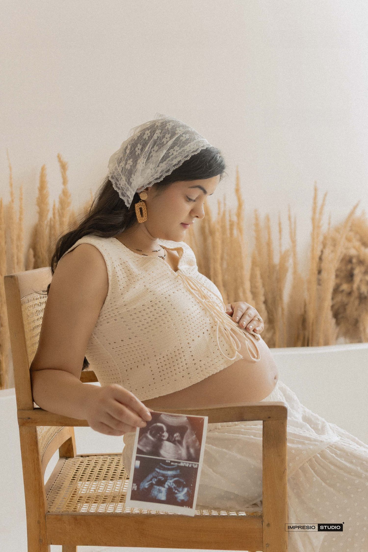 Pregnant woman in cream crochet top sits in wooden chair holding ultrasound photos, with dried pampas grass backdrop.