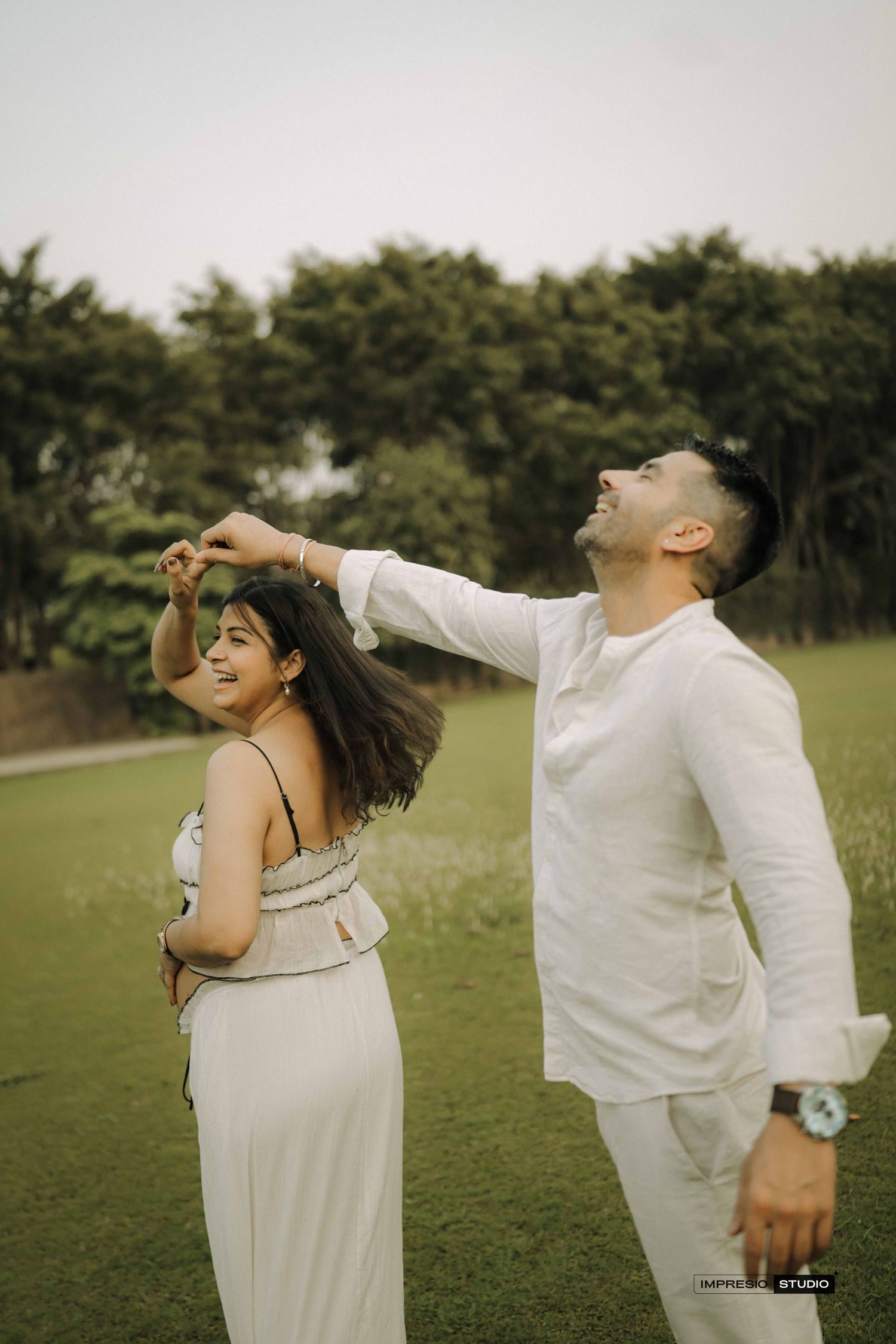 Couple in white outfits dancing outdoors on green grass with trees in background.
