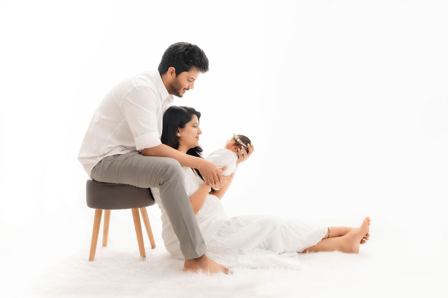 A couple in white clothing tenderly holds their newborn baby in a minimalist white studio setting.