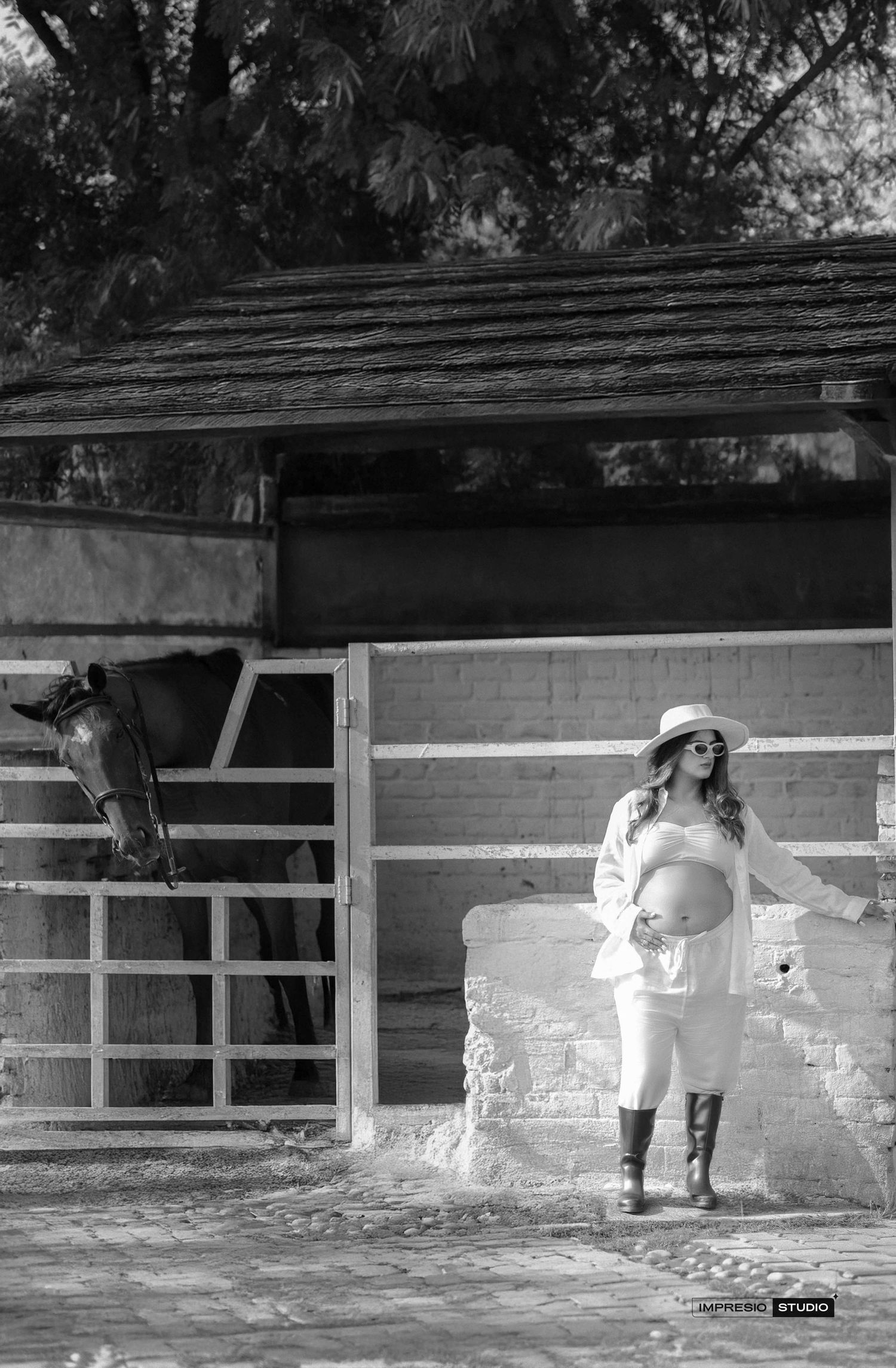 Black and white maternity photo of pregnant woman in hat and boots standing by rustic stable with horse.