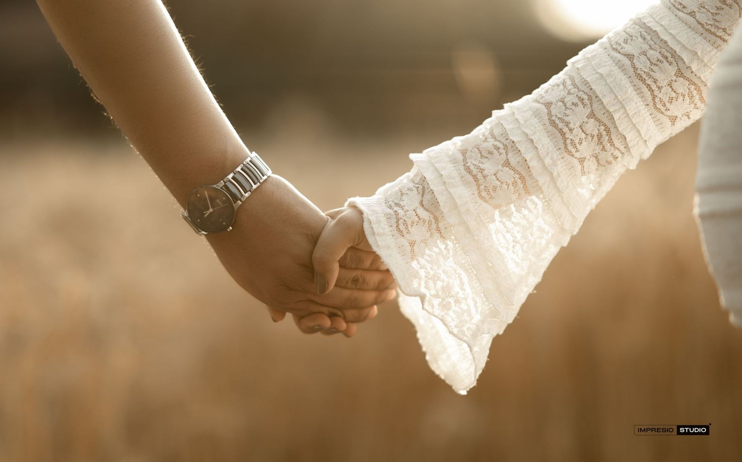 Two people holding hands in a sunlit field, one wearing a watch and the other in white lace, symbolizing love.