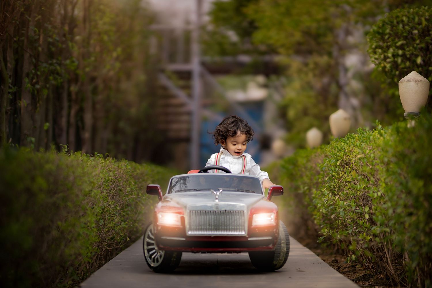 Toddler driving a toy Rolls-Royce electric car on a garden path surrounded by green hedges.
