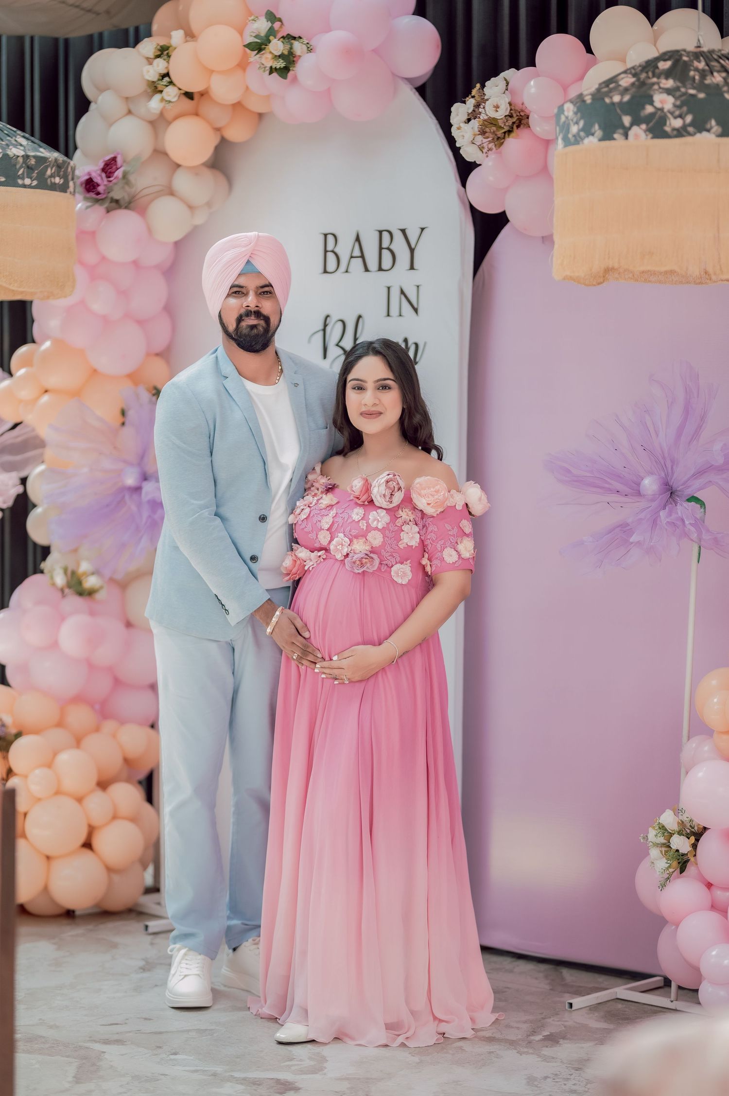 Couple at a baby shower, pregnant woman in pink floral gown, man in blue suit and turban, balloon arch backdrop.
