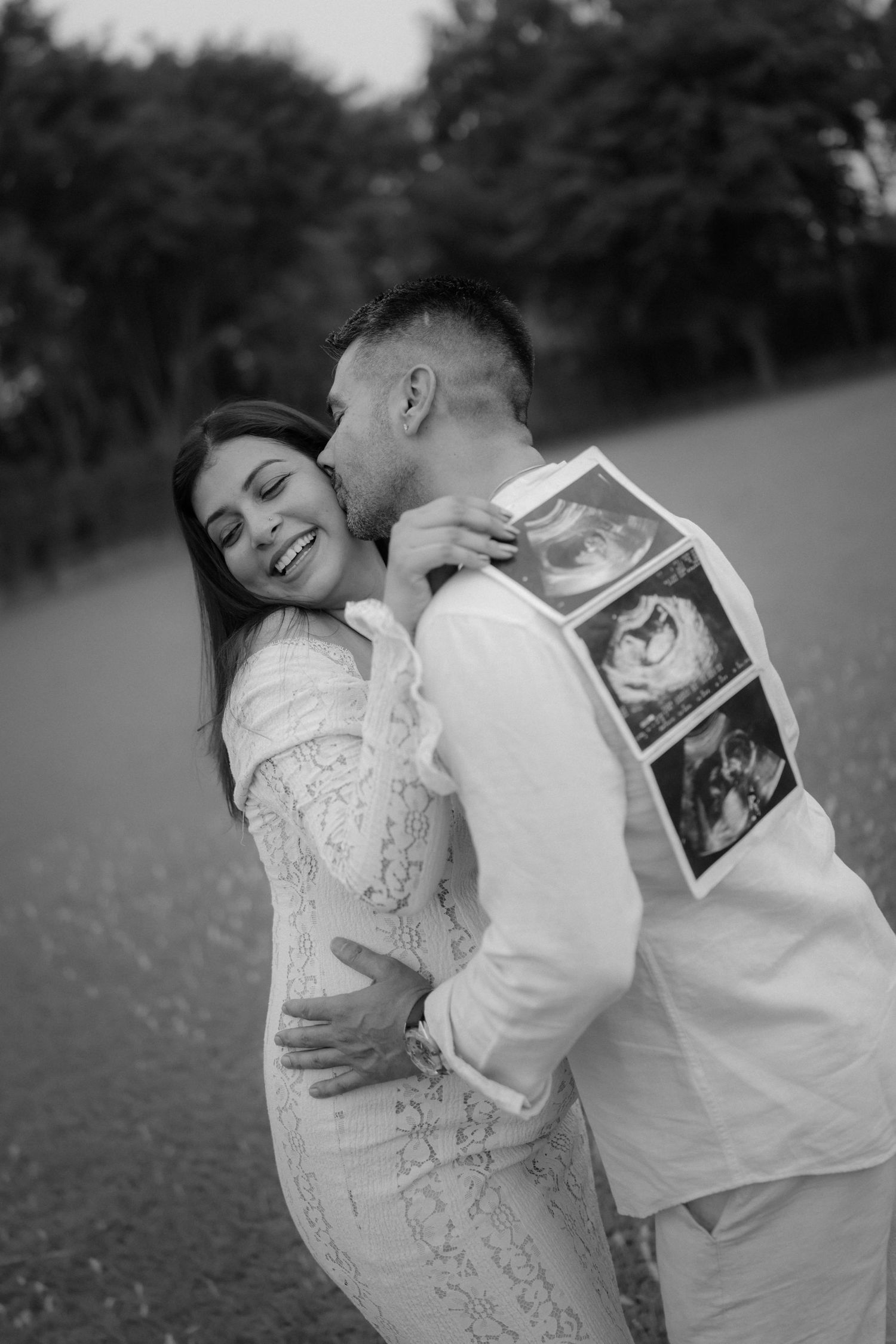 Black and white photo of a couple embracing outdoors, man kissing woman's cheek while holding ultrasound photos.