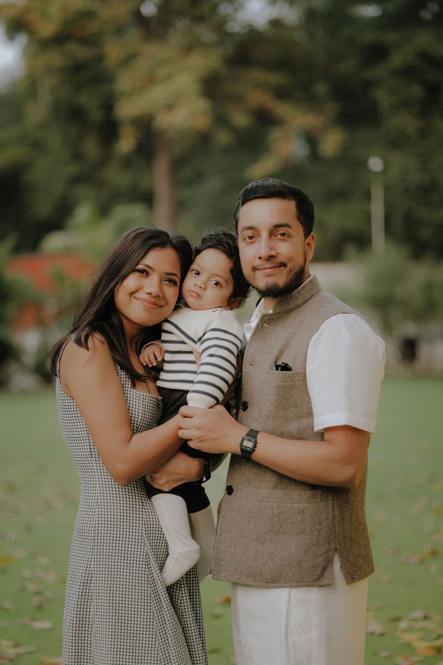 Happy young family with baby posing outdoors in a lush green park, wearing elegant traditional and casual attire.