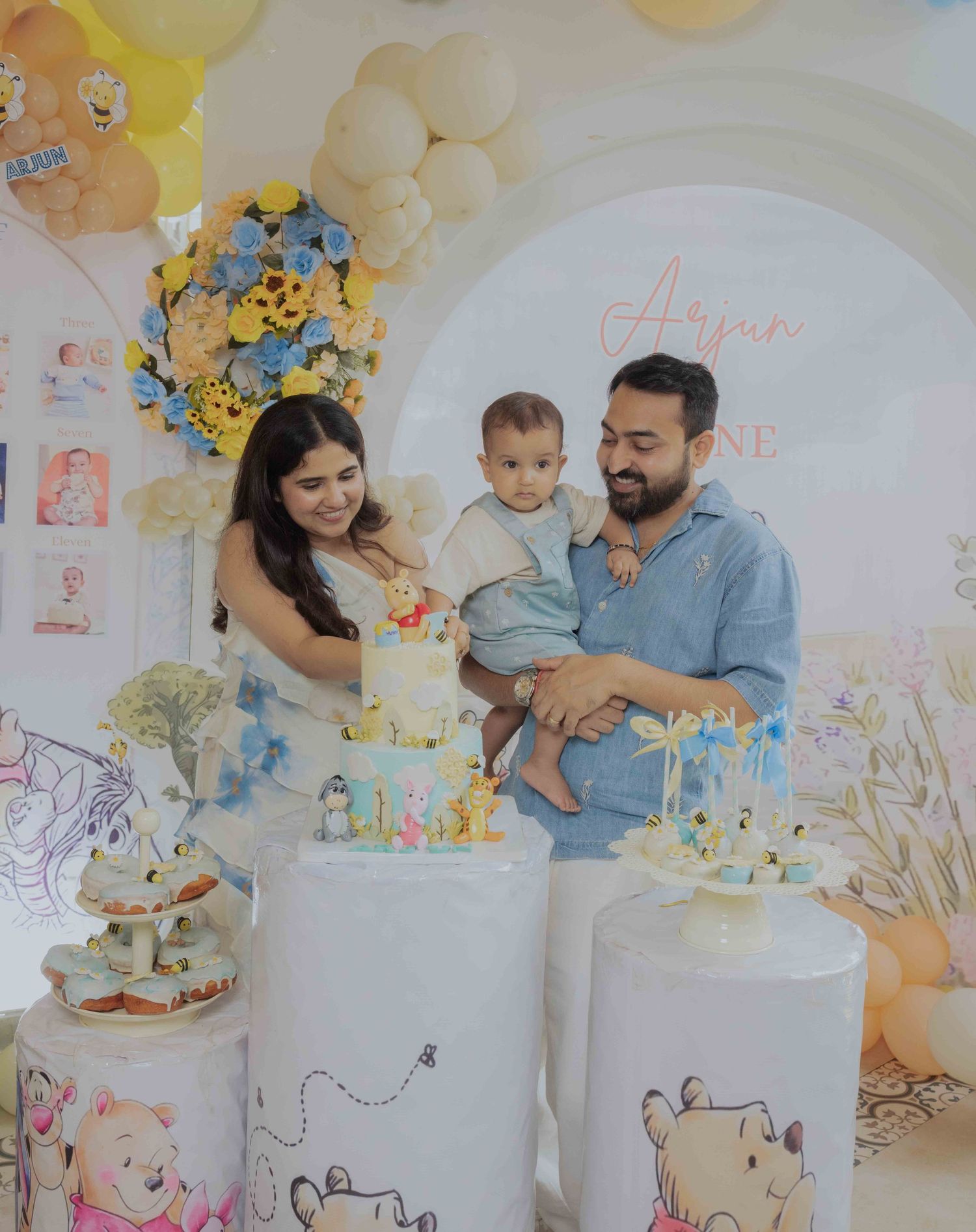 Family celebrating baby's first birthday with Winnie the Pooh themed cake and balloon decorations.