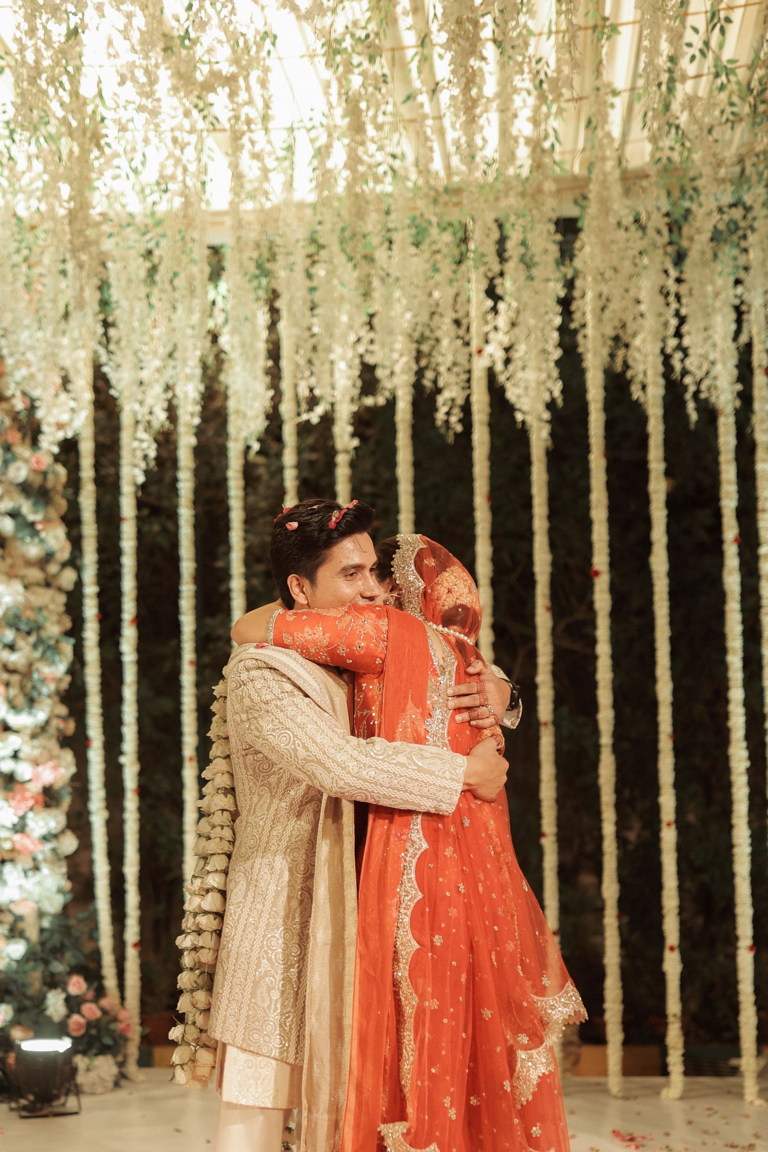 Groom embraces bride in red lehenga under elegant floral backdrop at Indian wedding.