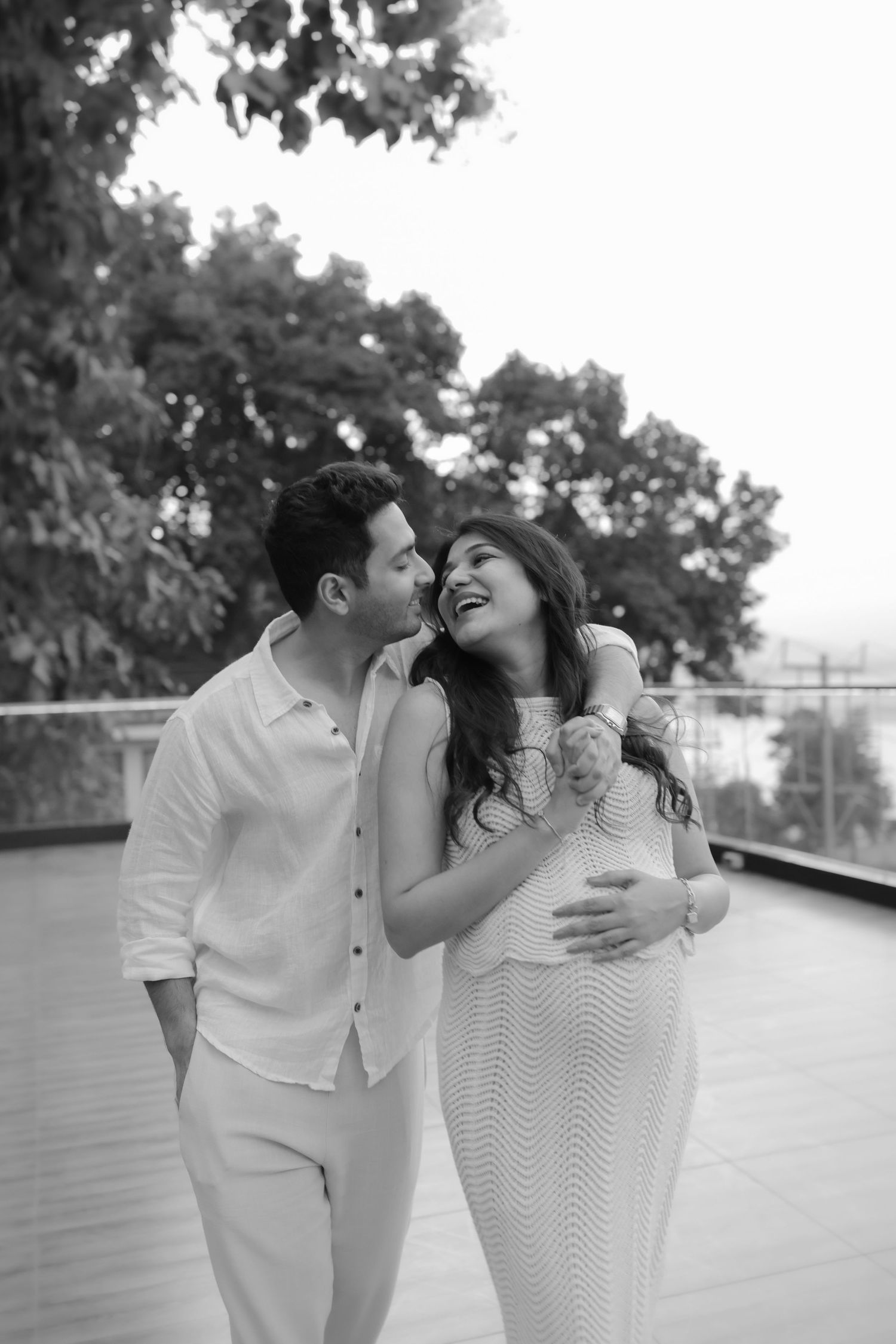 Black and white photo of a couple sharing a tender moment outdoors, woman in white dress cradling baby bump.