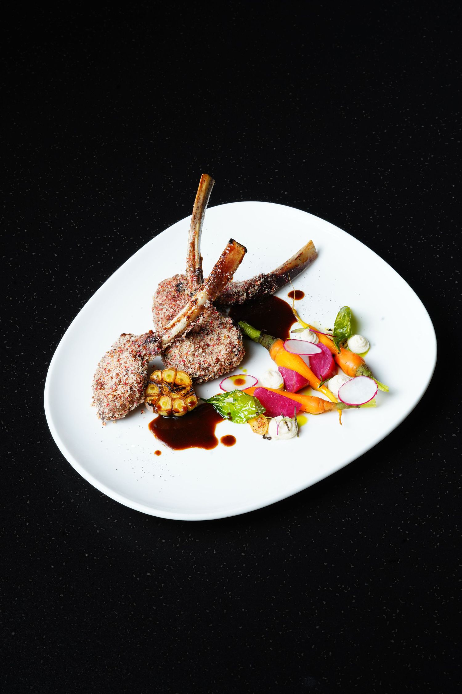 Elegant plated lamb chops with colorful vegetables and garnishes on white curved plate.