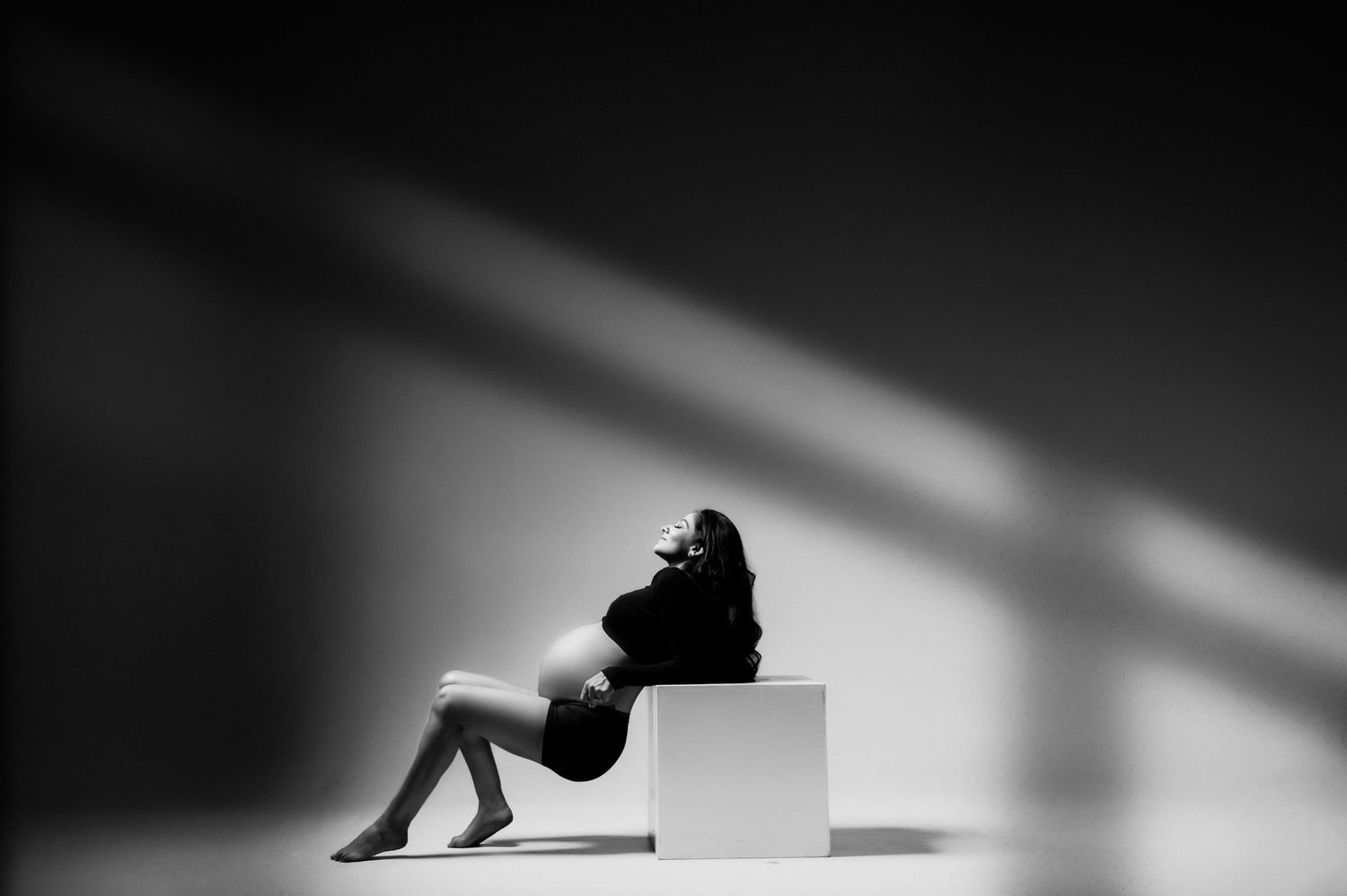 Black and white maternity photo of pregnant woman sitting on white cube with dramatic shadows.