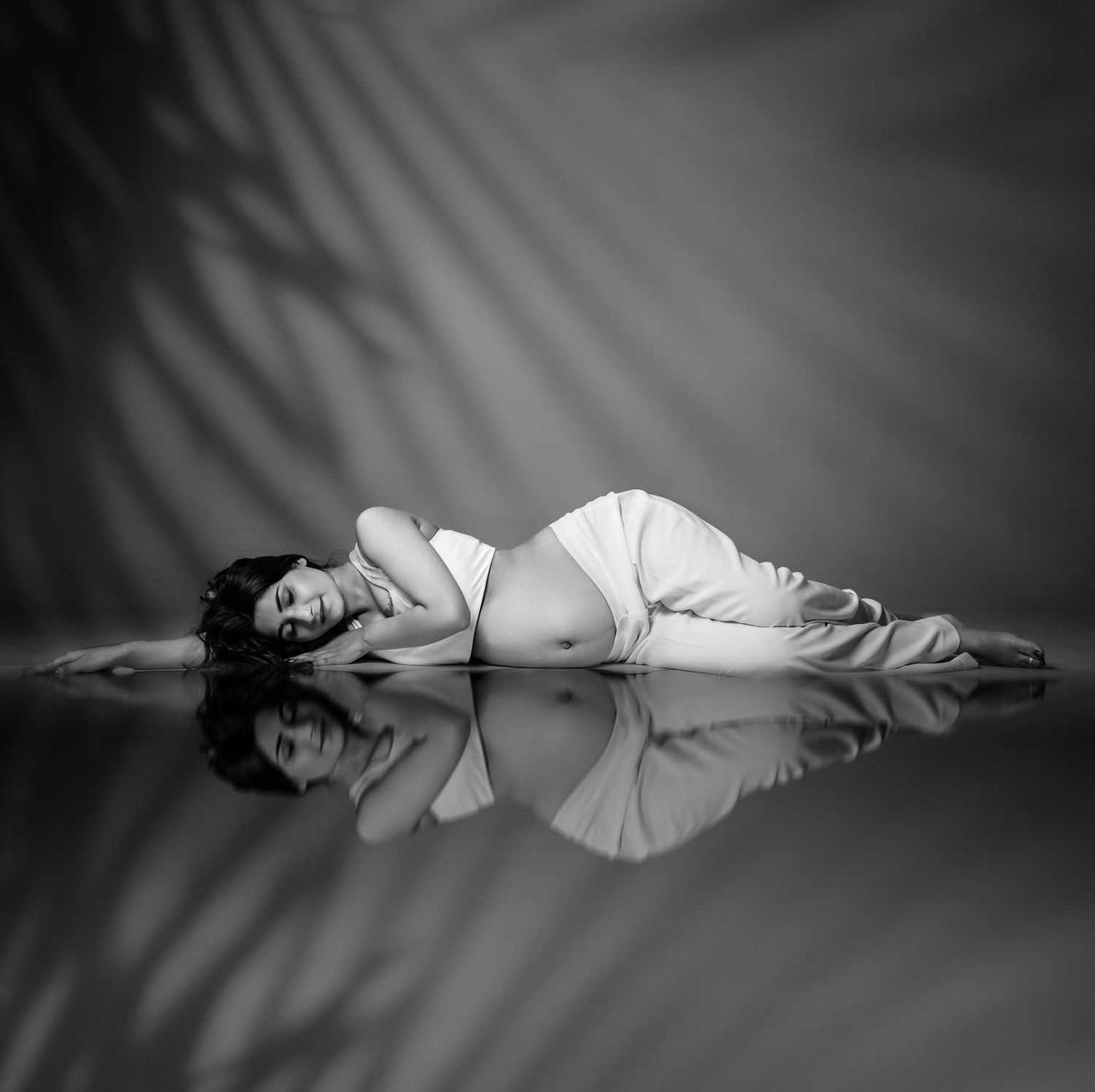 Black and white photo of a pregnant woman lying on a reflective floor with palm leaf shadows in the background.