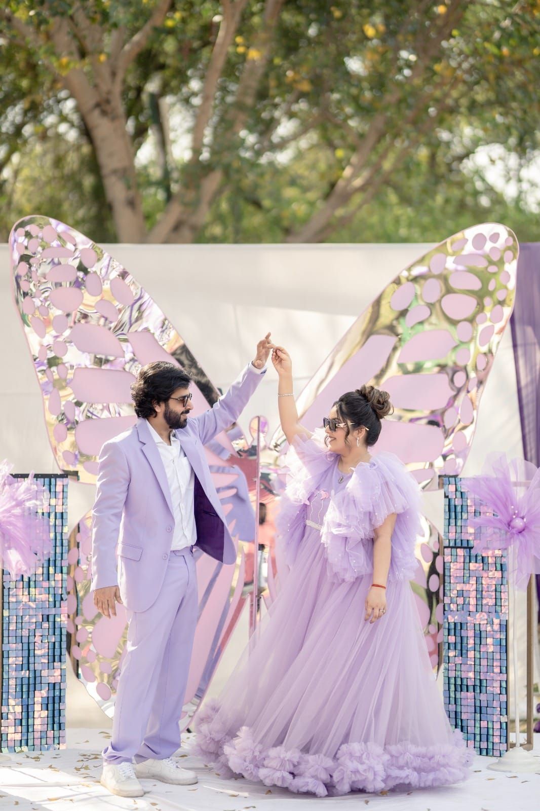 Couple in matching lavender outfits posing before a large decorative butterfly backdrop at an outdoor event.
