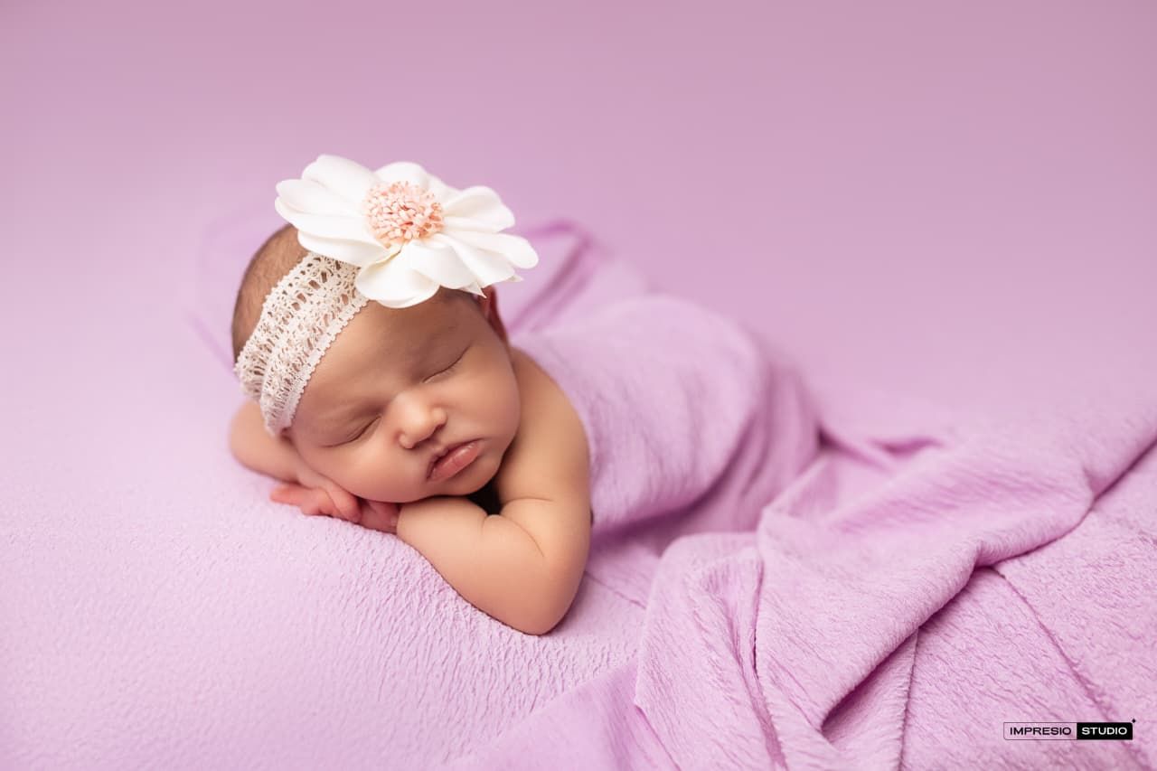 Sleeping newborn baby girl wrapped in pink blanket with floral headband on lavender background.
