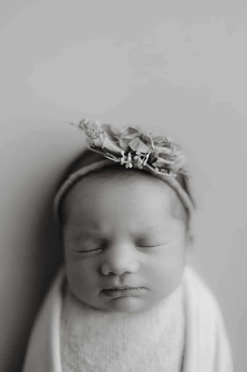 Black and white newborn portrait of a sleeping baby girl wearing a delicate floral crown, wrapped in soft white fabric.