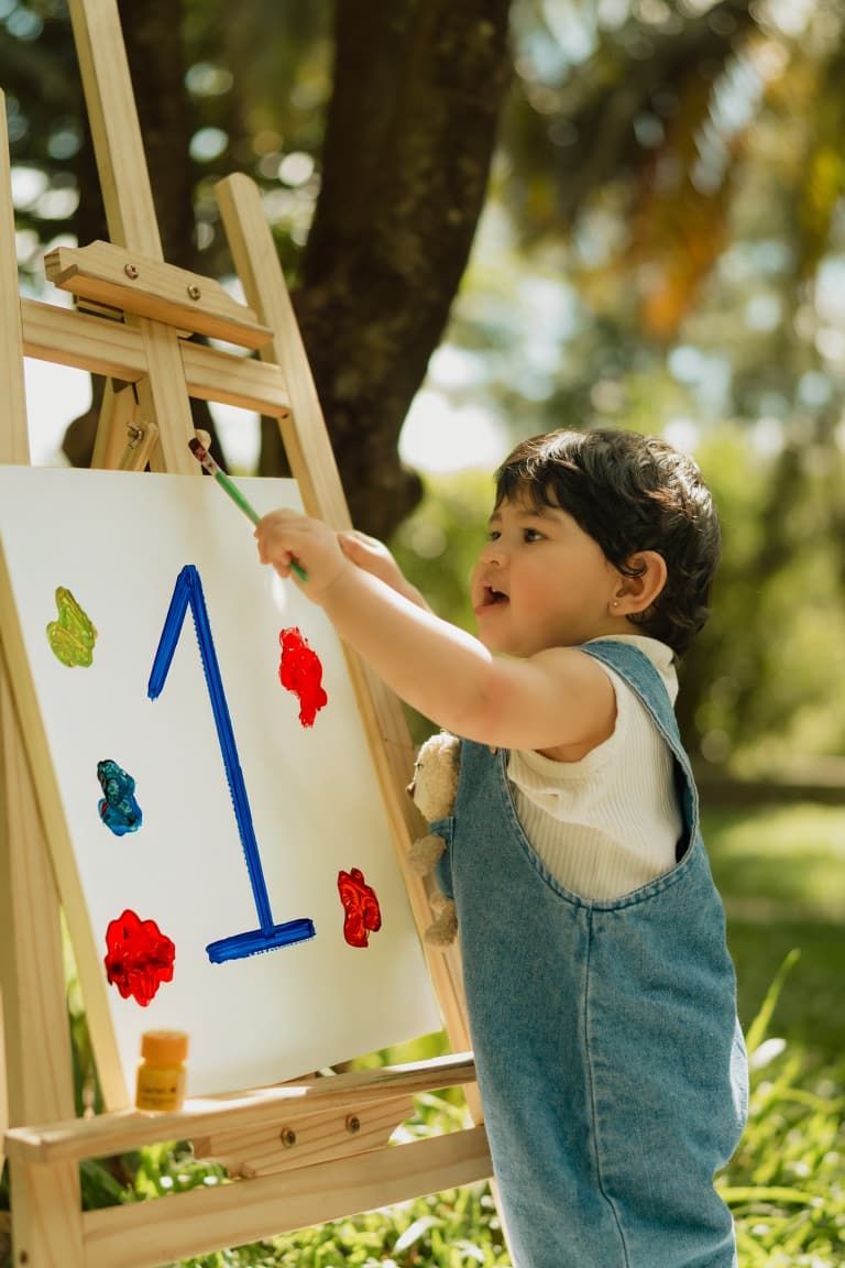 Toddler in denim overalls painting a colorful canvas on a wooden easel outdoors in a green park.