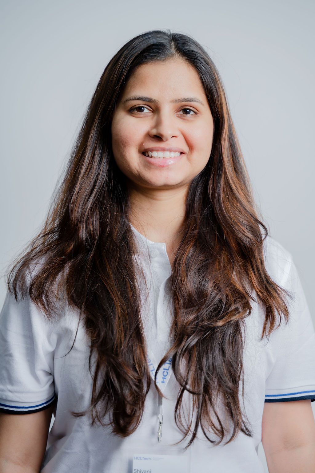 Smiling young woman with long brown hair wearing a white uniform top with blue trim against a gray background.
