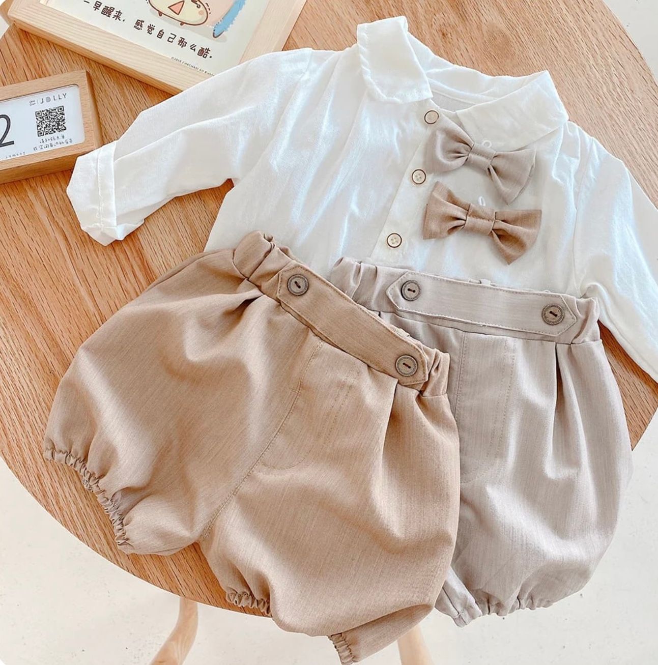 A set of beige and white baby romper outfits with coordinating bows laid out on a wooden floor.