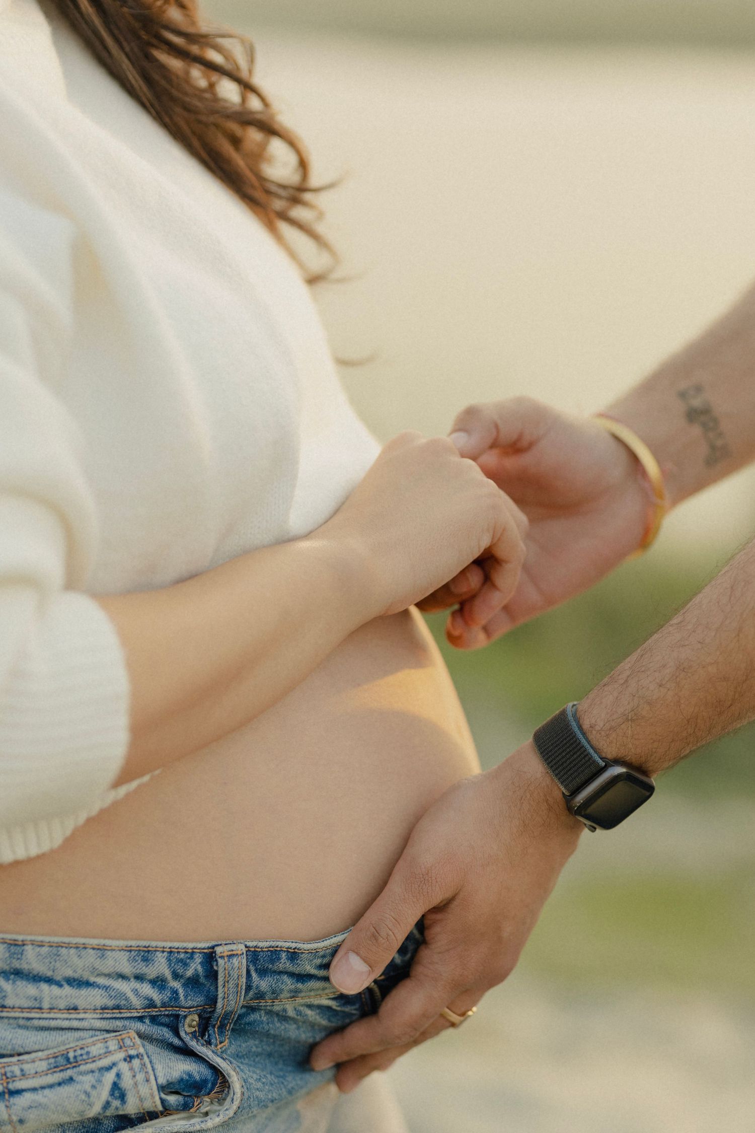 A man gently holds a pregnant woman's baby bump while she holds his hand outdoors in soft golden light.
